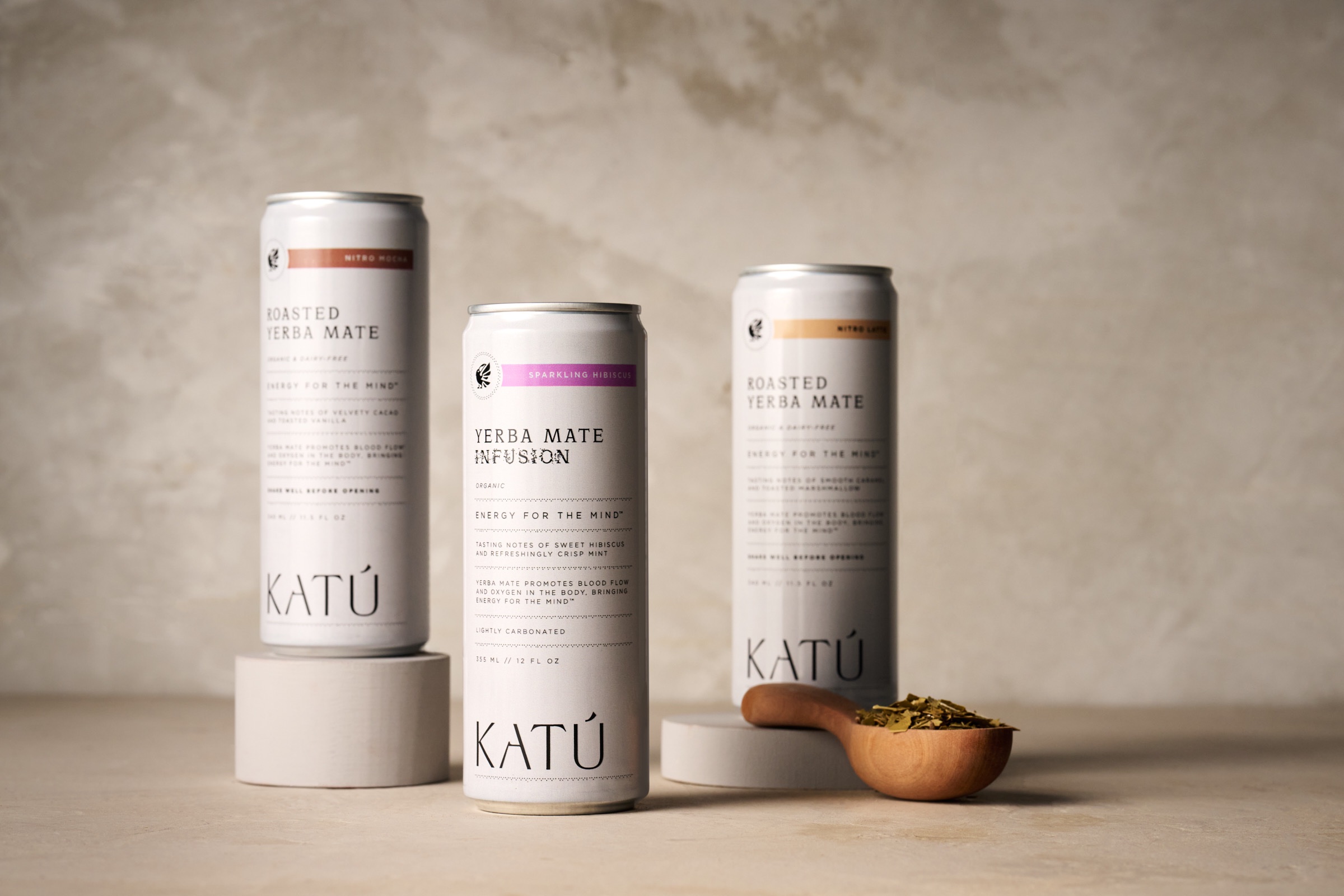 Three cans of KATÚ roasted yerba mate displayed on a neutral background, with a wooden scoop of loose yerba mate beside them.