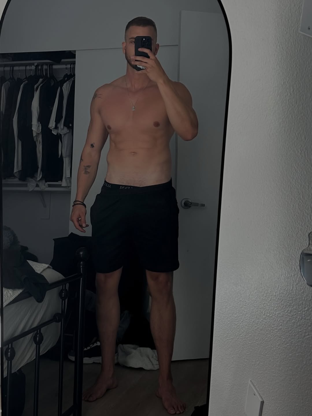 A person standing in front of a mirror, showing their upper body without a shirt, wearing black shorts. The background includes a closet and a bed.