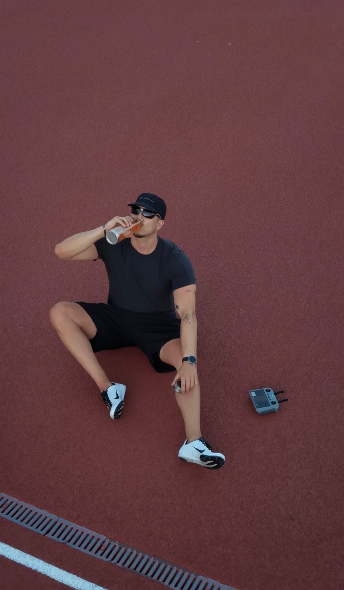 A person sitting on a red athletic track, sipping from a can, wearing a casual outfit and sneakers.