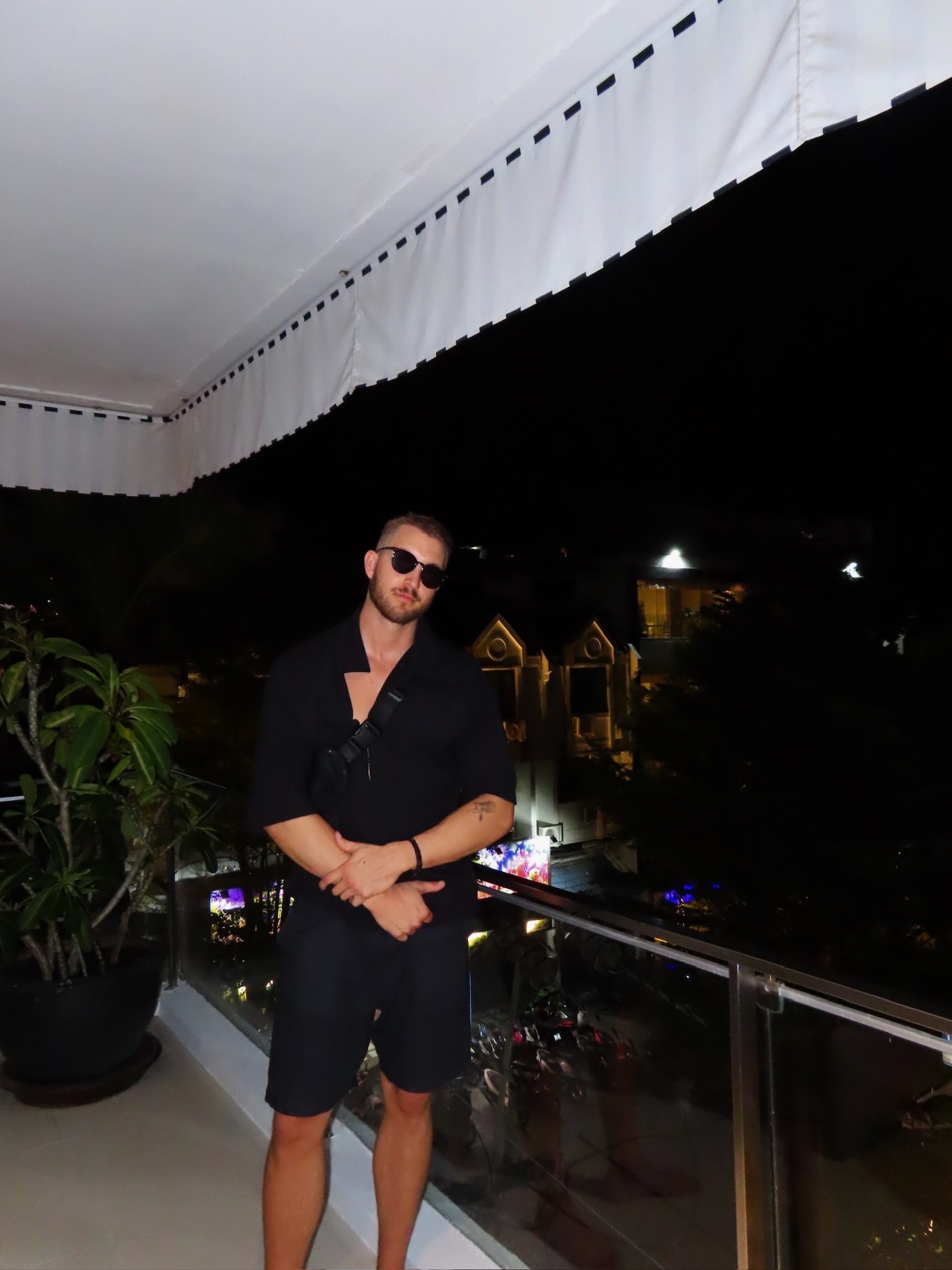 A man wearing sunglasses and a black outfit stands on a balcony at night, with a cityscape visible in the background.