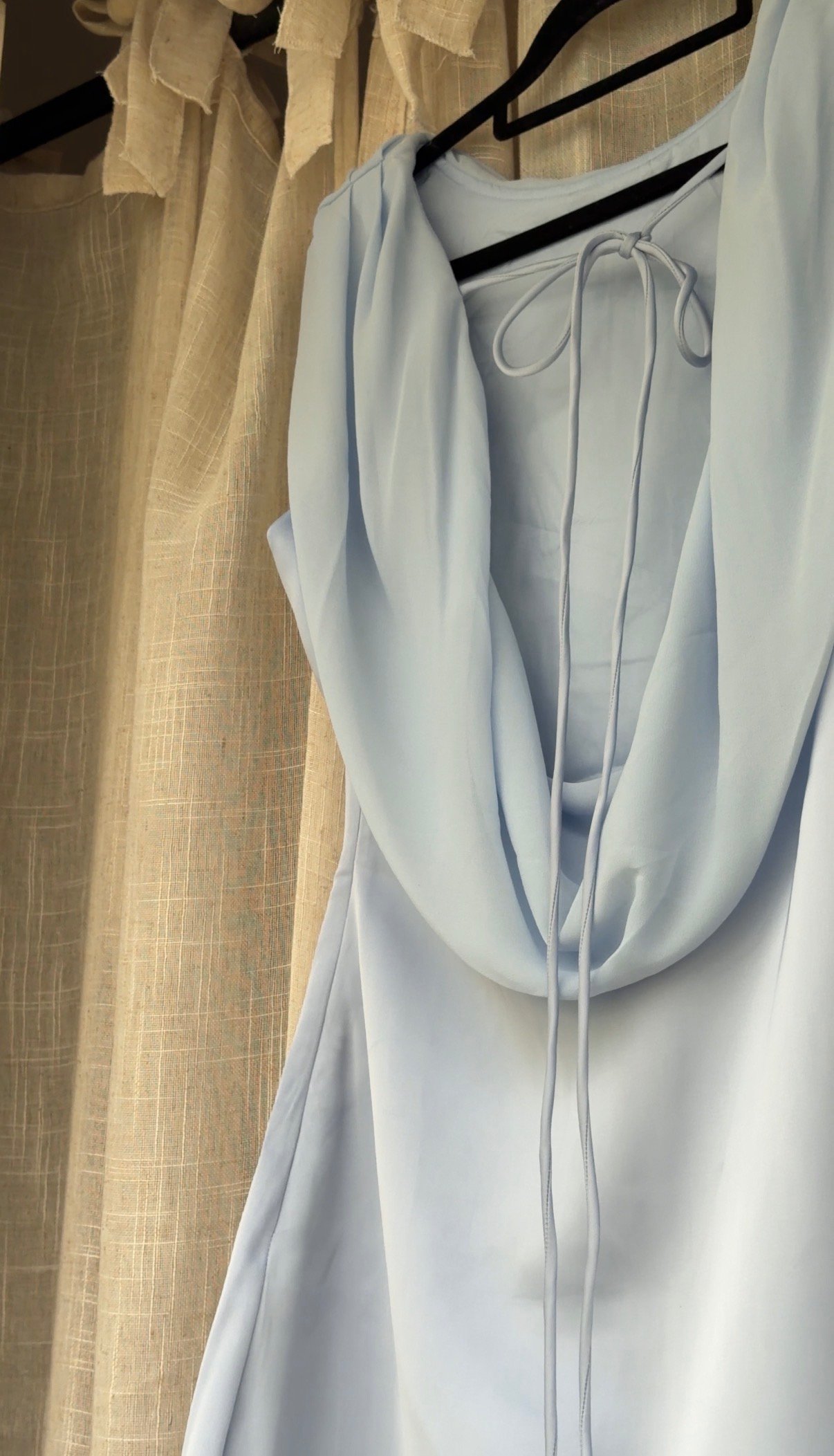 A light blue dress hanging on a hanger, with soft draping and a tied detail at the neckline, set against a textured curtain backdrop.