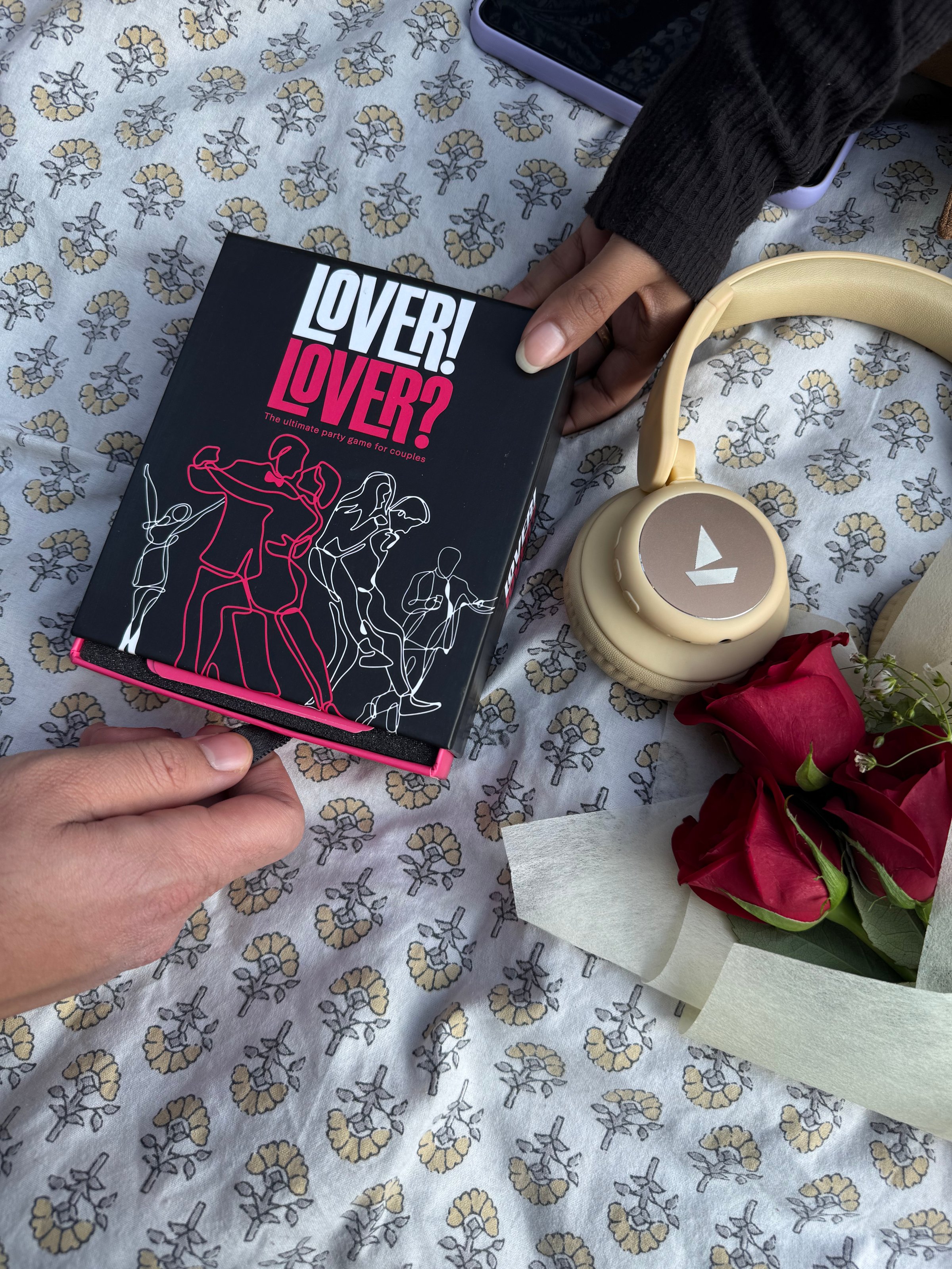A close-up of a book titled 'Lover! Lover?' held by a hand, alongside a pair of headphones and a bouquet of red roses on a patterned surface.