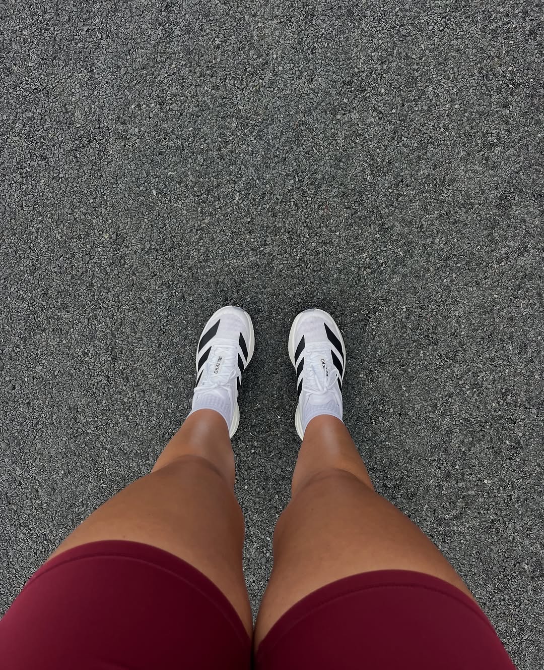 A close-up view of a person's legs wearing maroon shorts and white athletic shoes on a textured gray surface.