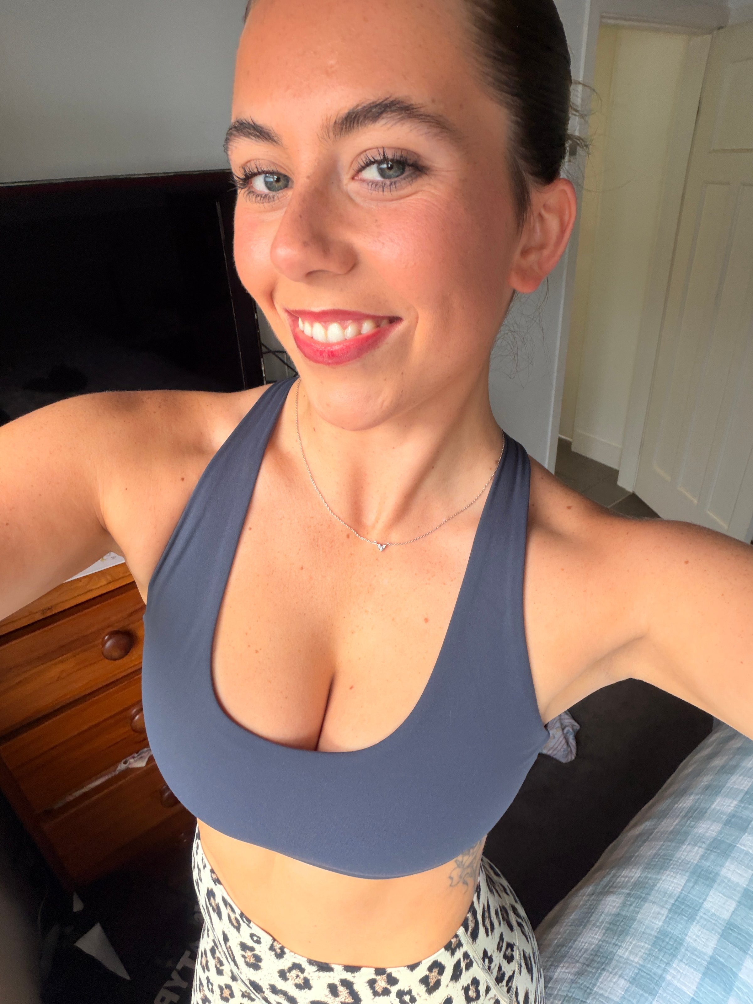 A young woman smiling and posing for a selfie, wearing a dark sports bra and patterned leggings, with a cozy room in the background.