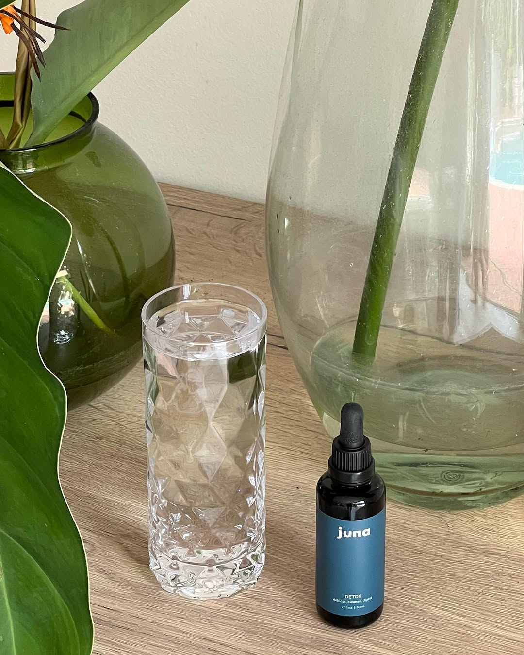 A clear glass of water sits next to a small bottle of detox oil on a wooden surface, with green plants and a large glass vase in the background.