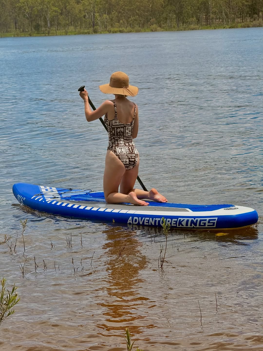 A person in a patterned swimsuit paddles on a blue stand-up paddleboard in calm water, wearing a wide-brimmed hat.