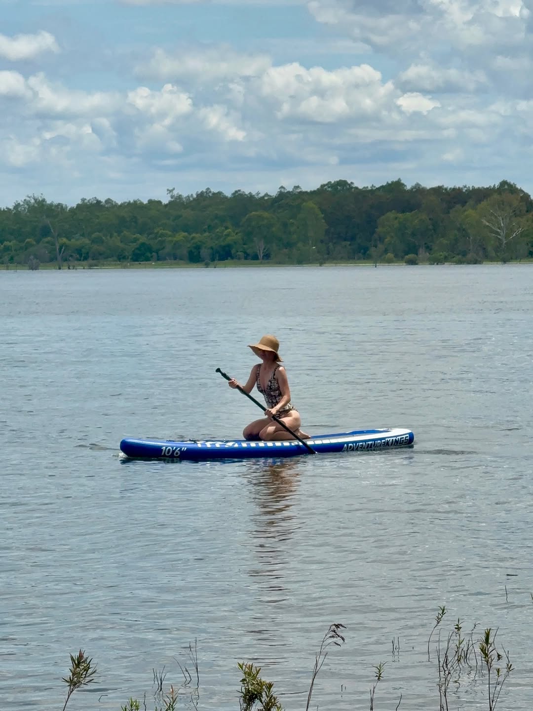 A person paddles on a stand-up paddleboard in a calm lake surrounded by greenery under a partly cloudy sky.