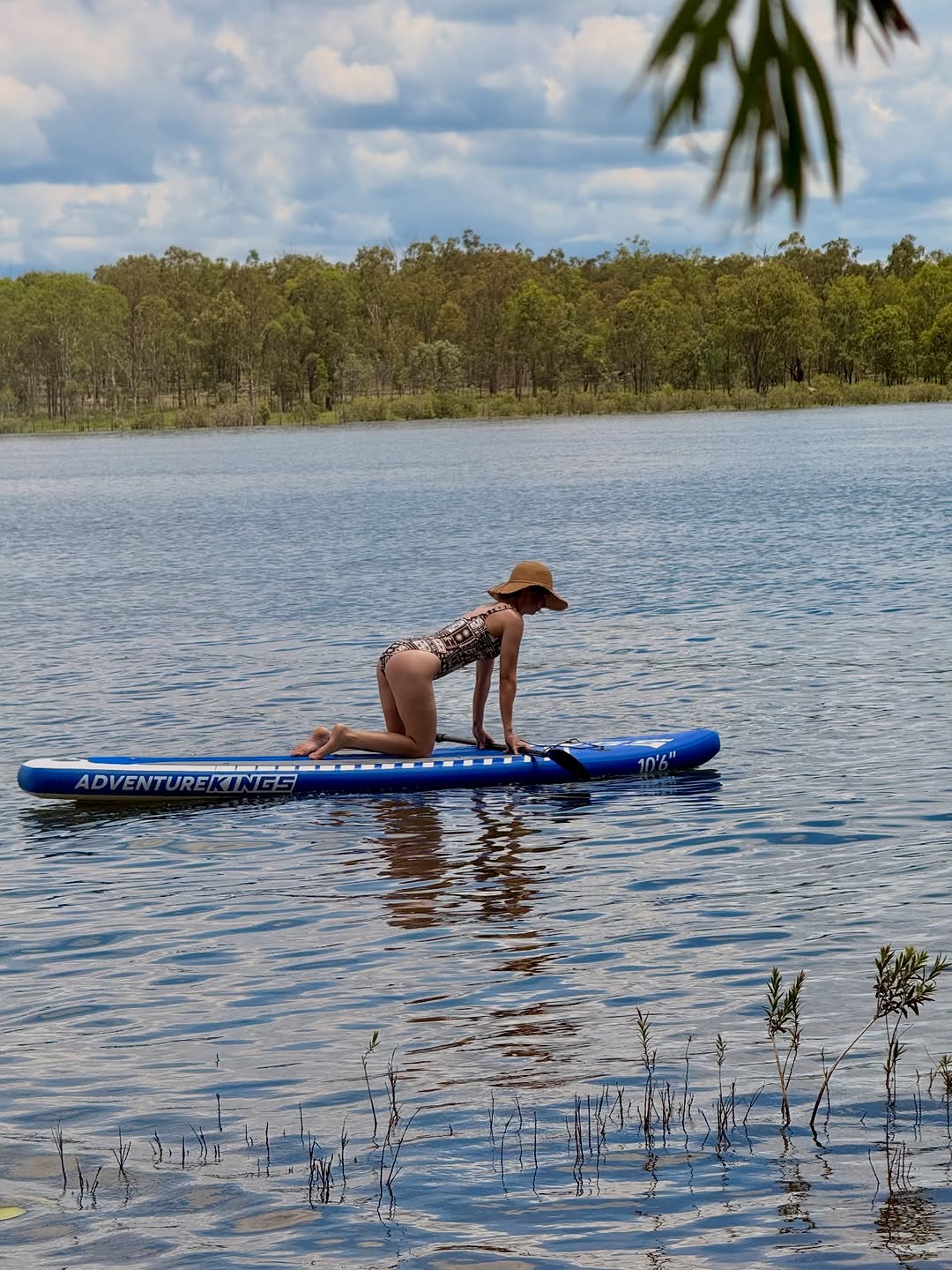 A person in a swimsuit kneels on a blue paddleboard in a calm lake surrounded by greenery under a partly cloudy sky.