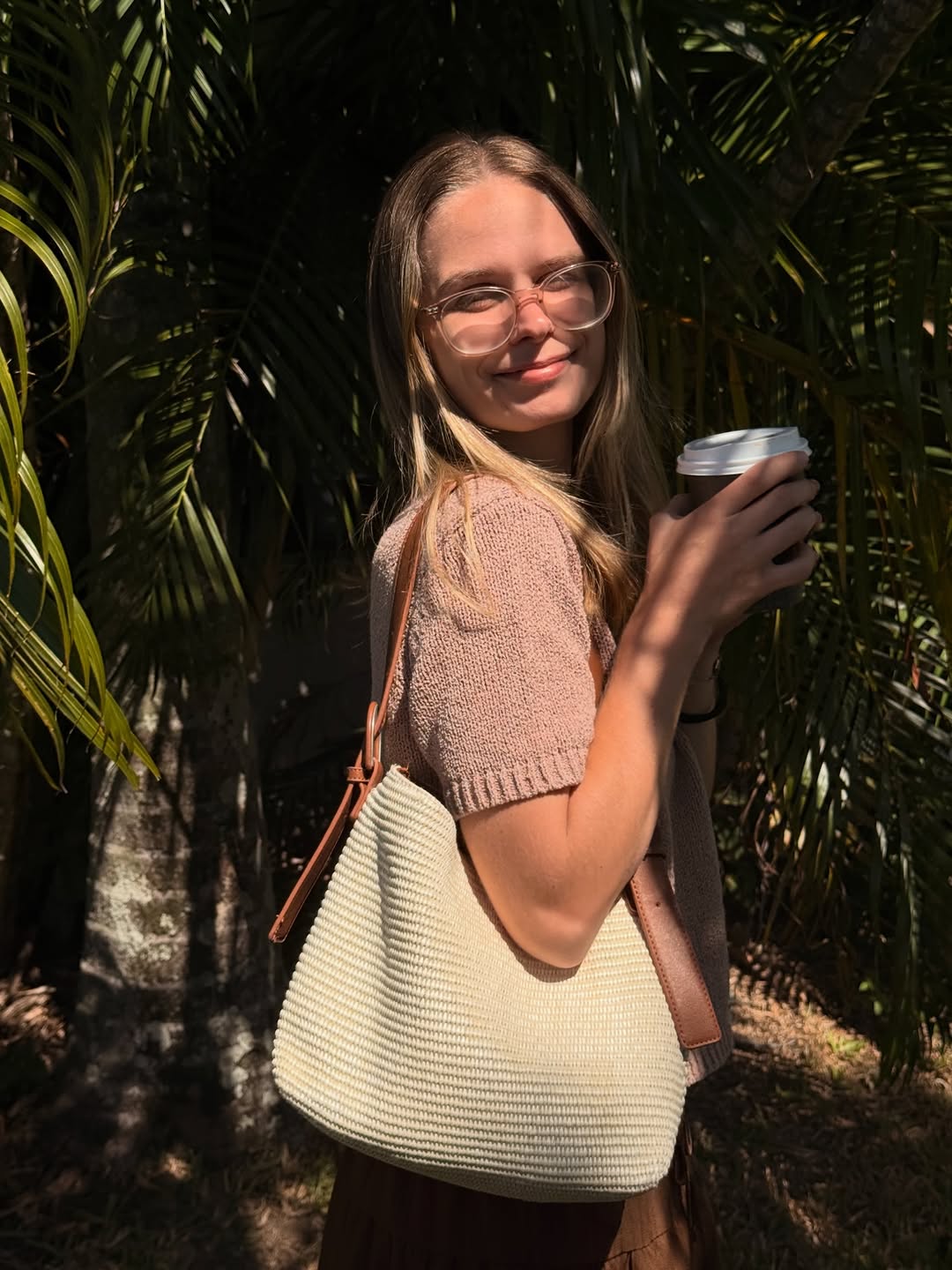 A woman stands outdoors among palm leaves, holding a coffee cup and wearing a stylish knitted sweater and a light-colored handbag.