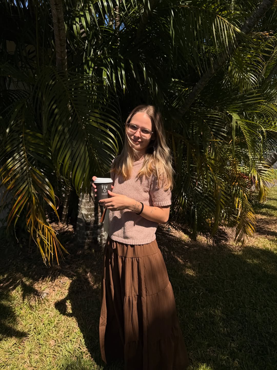 A young woman stands outdoors in front of lush green palm leaves, wearing a light sweater and a long brown skirt, holding a coffee cup.