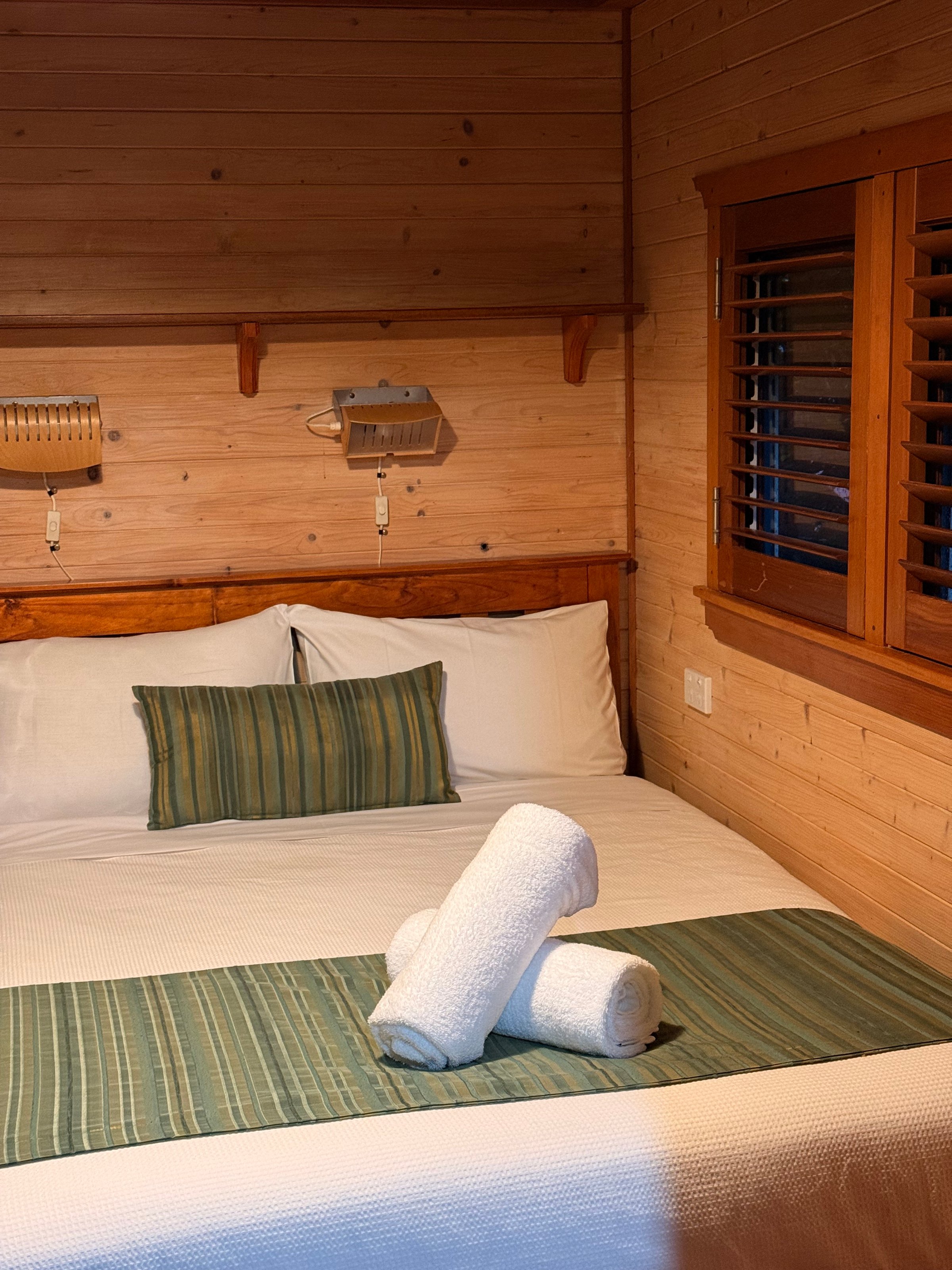 A cozy wooden bedroom featuring a neatly made bed with white linens and a green striped throw. Rolled towels are placed decoratively on the bed.