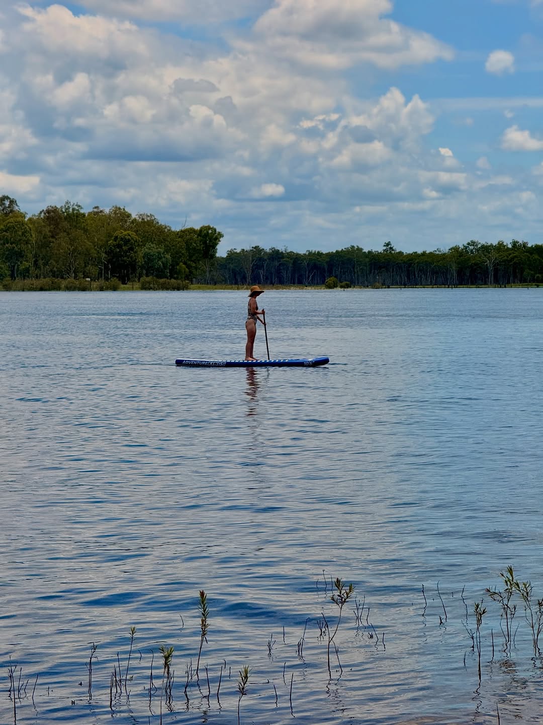 A person stands on a paddleboard in a calm lake surrounded by trees and a cloudy sky.