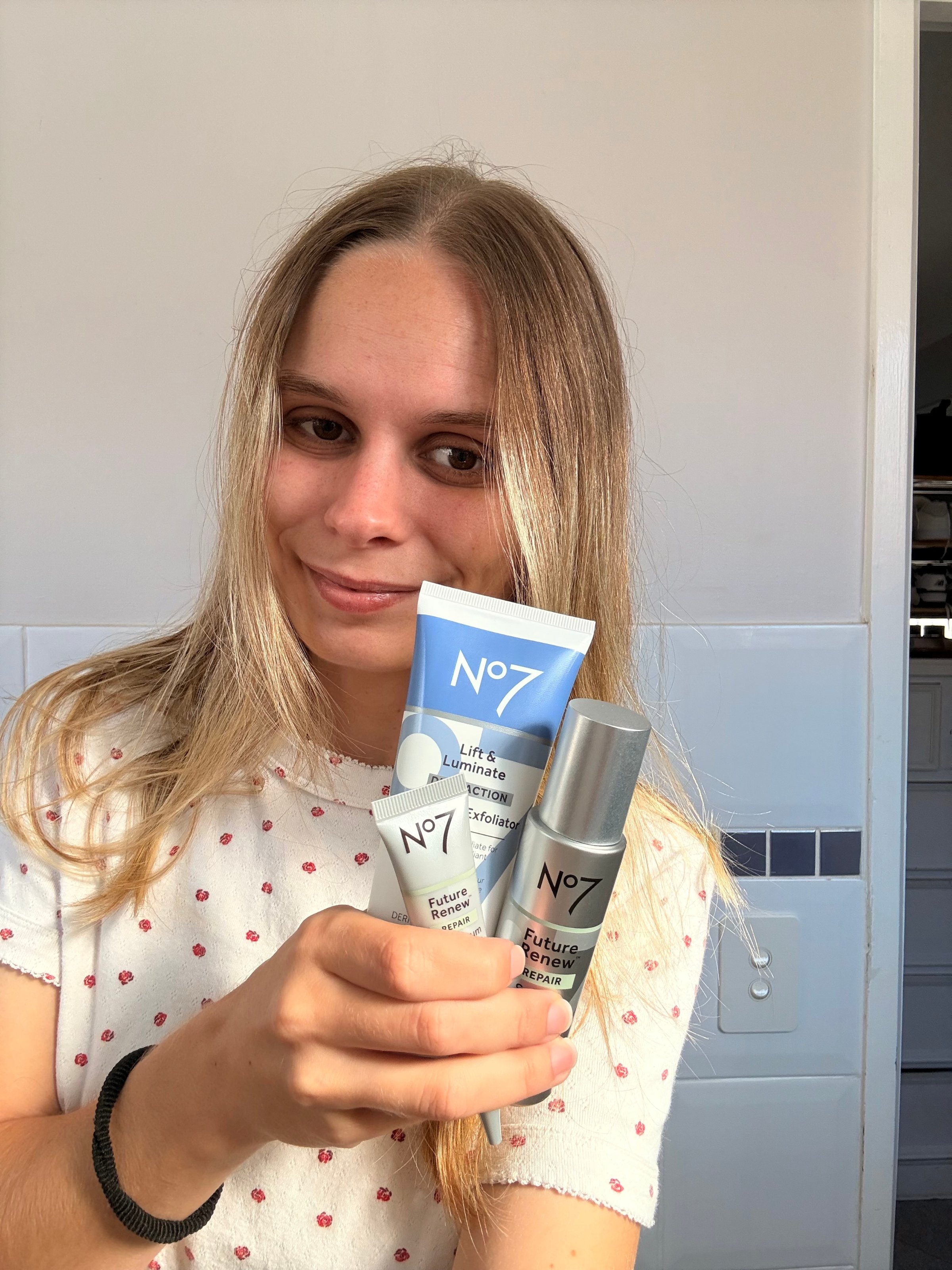 A person holding three skincare products from the No7 brand in a bathroom setting.