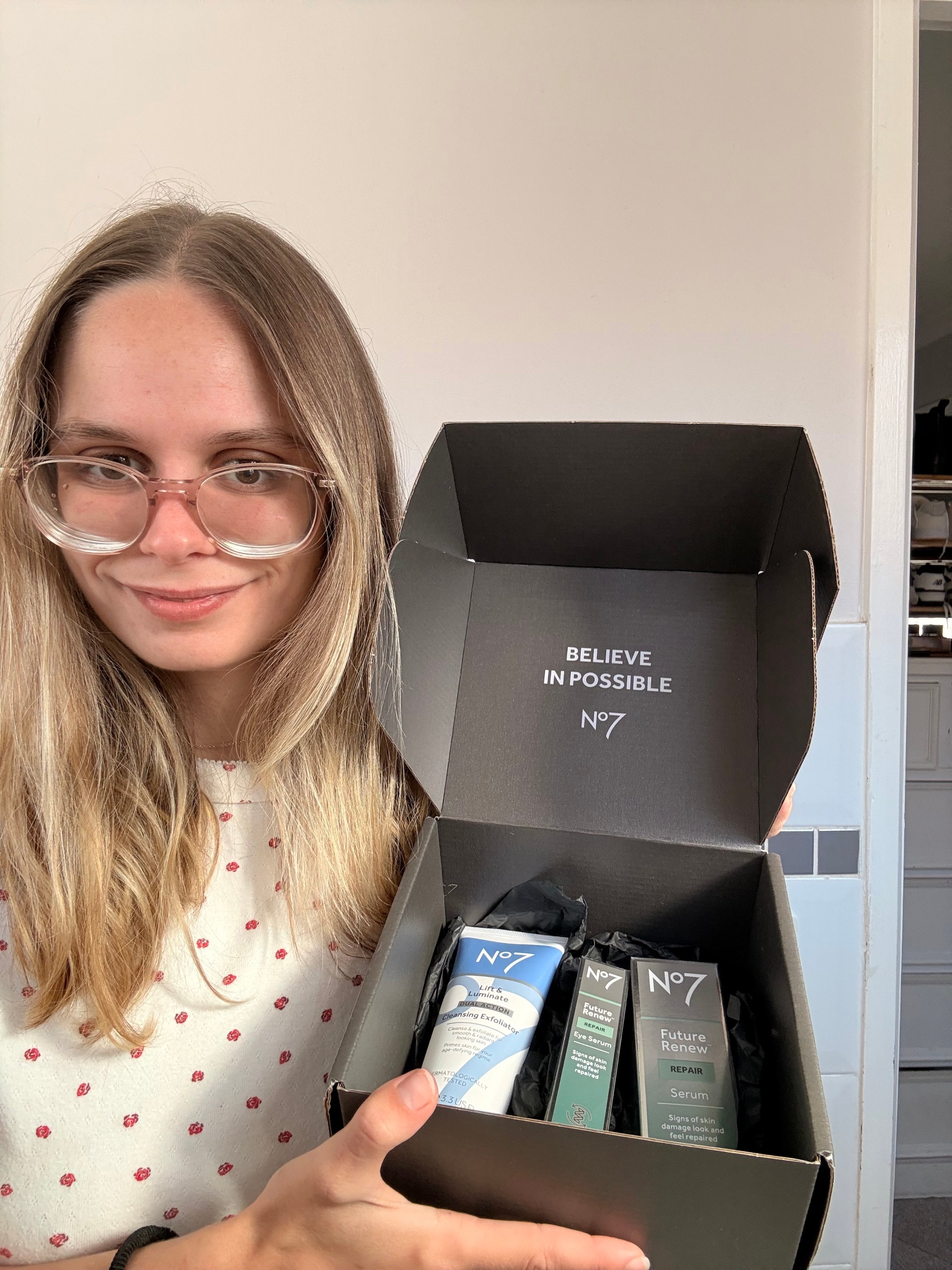 A young woman with long hair and glasses holds a black box containing skincare products, with the text 'BELIEVE IN POSSIBLE' visible inside the box.