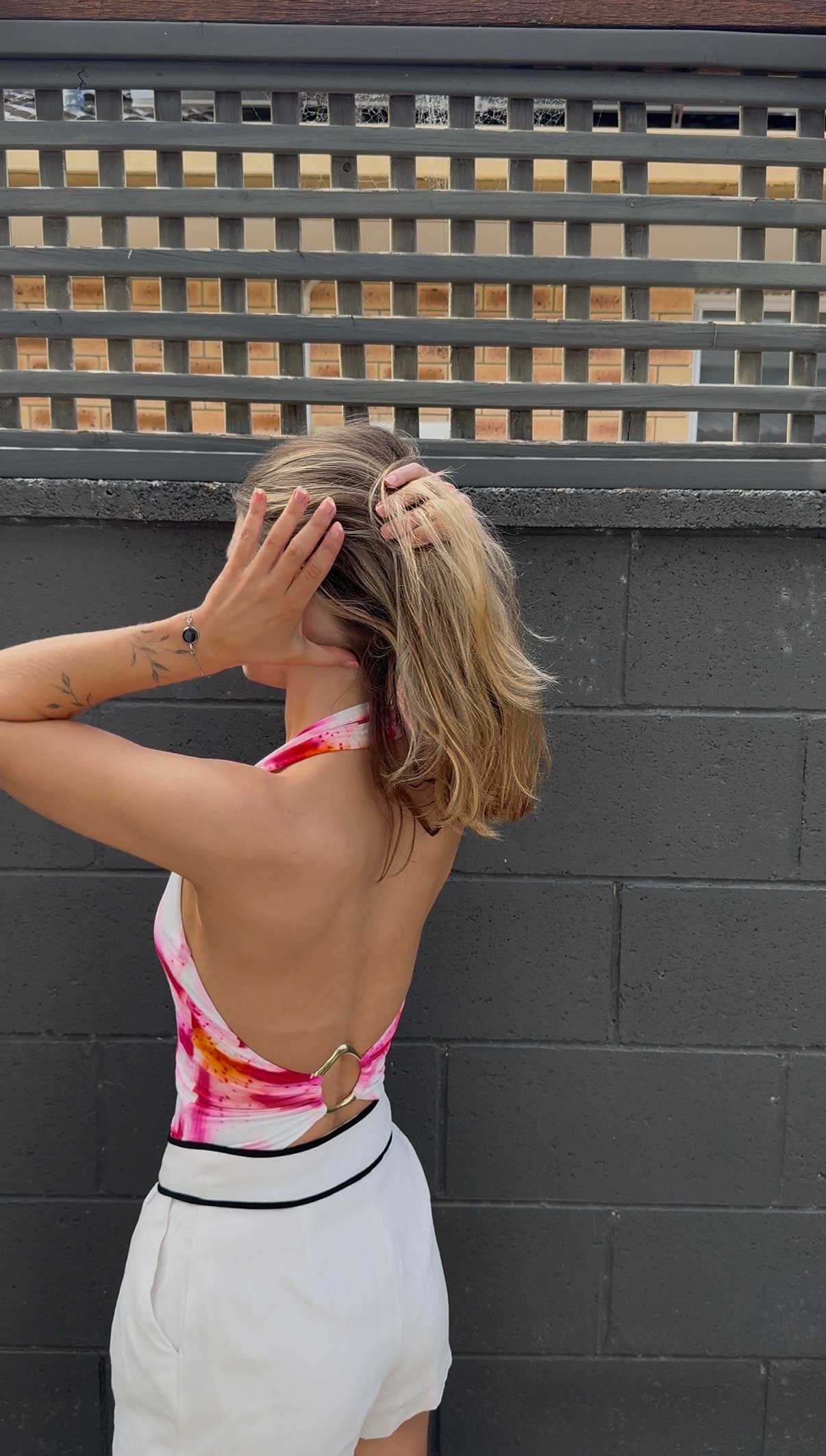 A woman with long hair styled in a casual manner, wearing a colorful halter top and white shorts, stands with her back to the camera against a textured wall.
