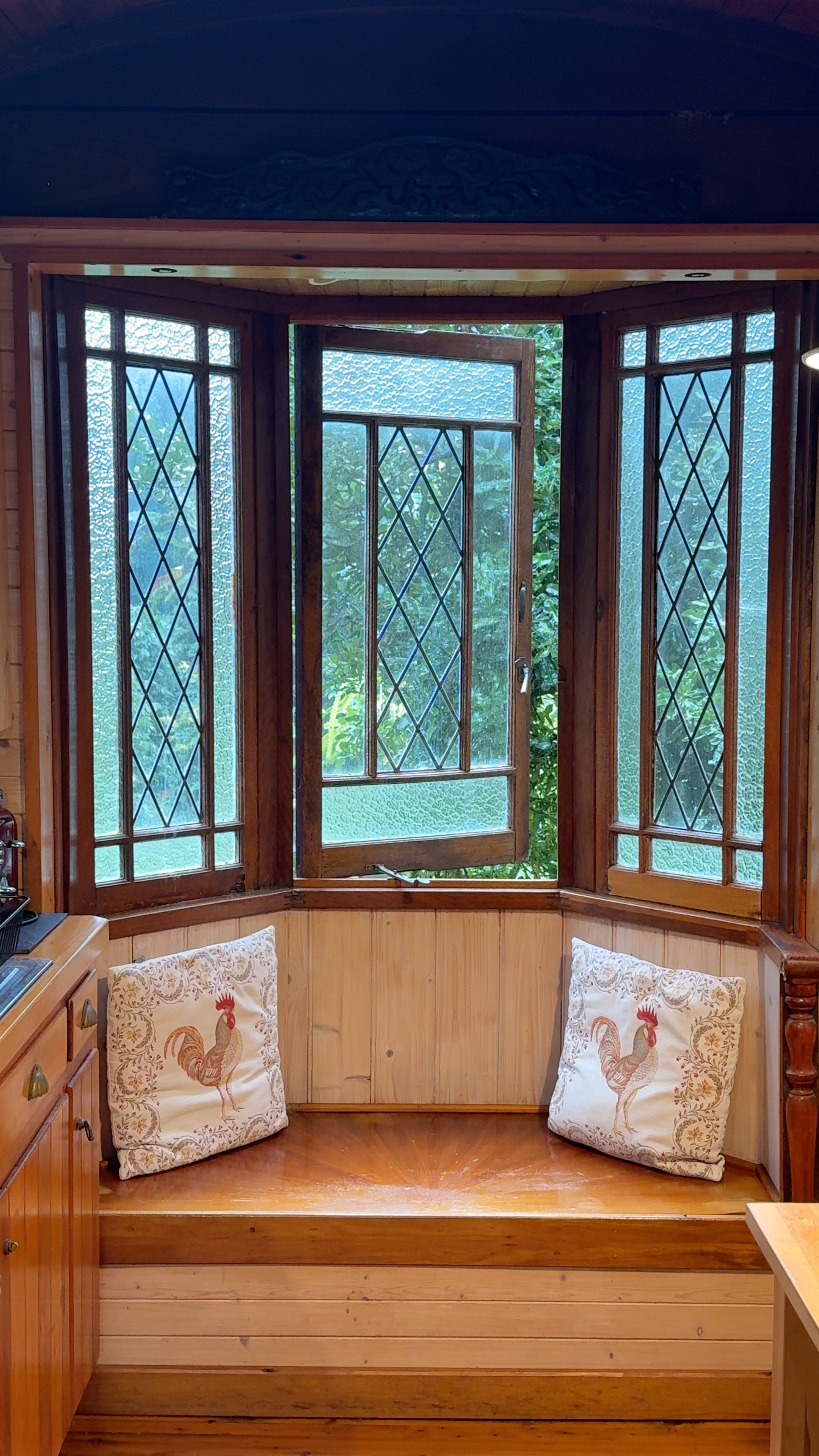 A cozy kitchen nook with a wooden bench and decorative pillows, featuring large diamond-patterned windows that let in natural light and a view of greenery outside.