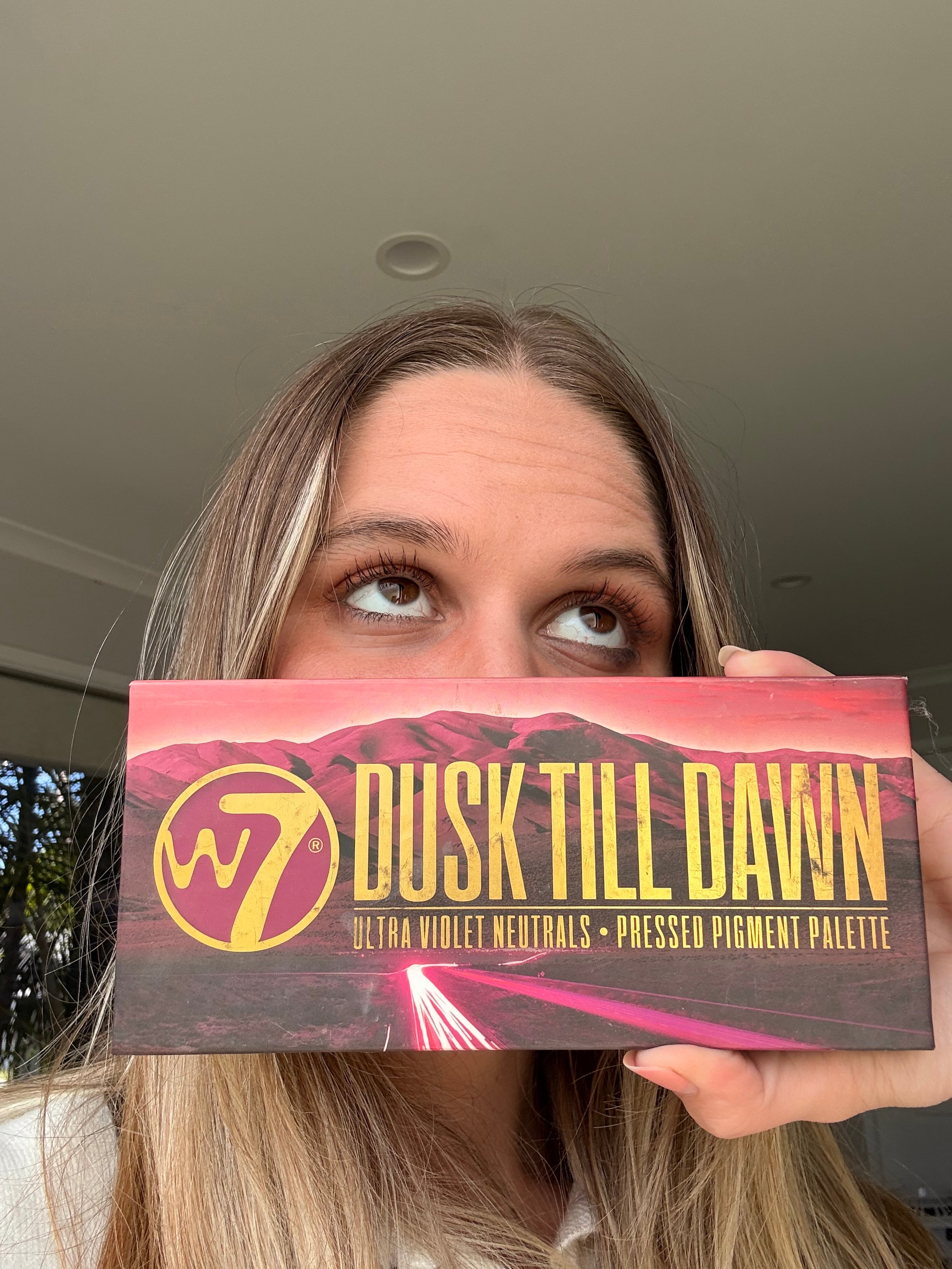 A person holds a colorful eyeshadow palette titled 'Dusk Till Dawn' close to their face, with a blurred background.