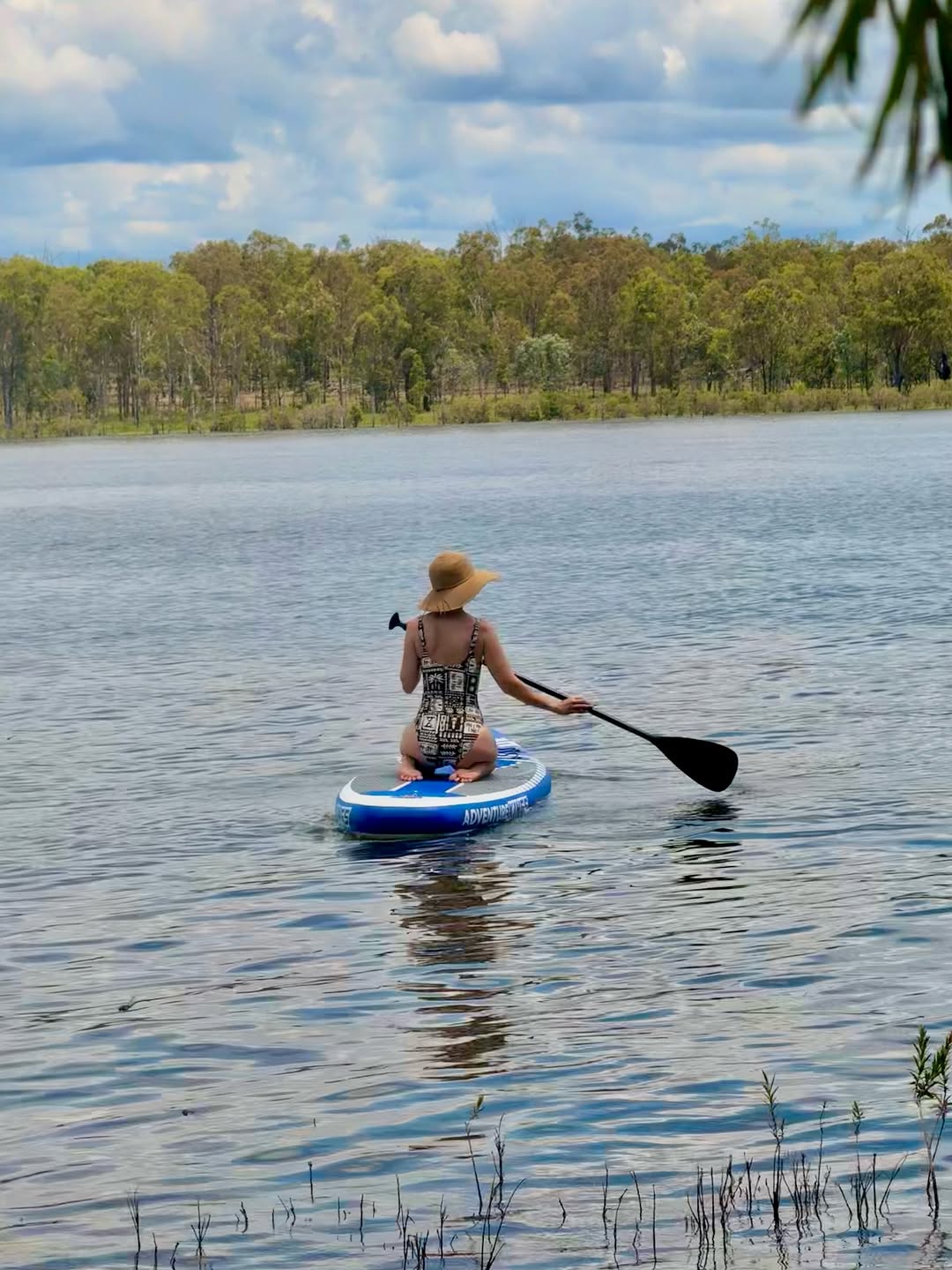 A person paddles on a stand-up paddleboard in a serene body of water surrounded by lush greenery.