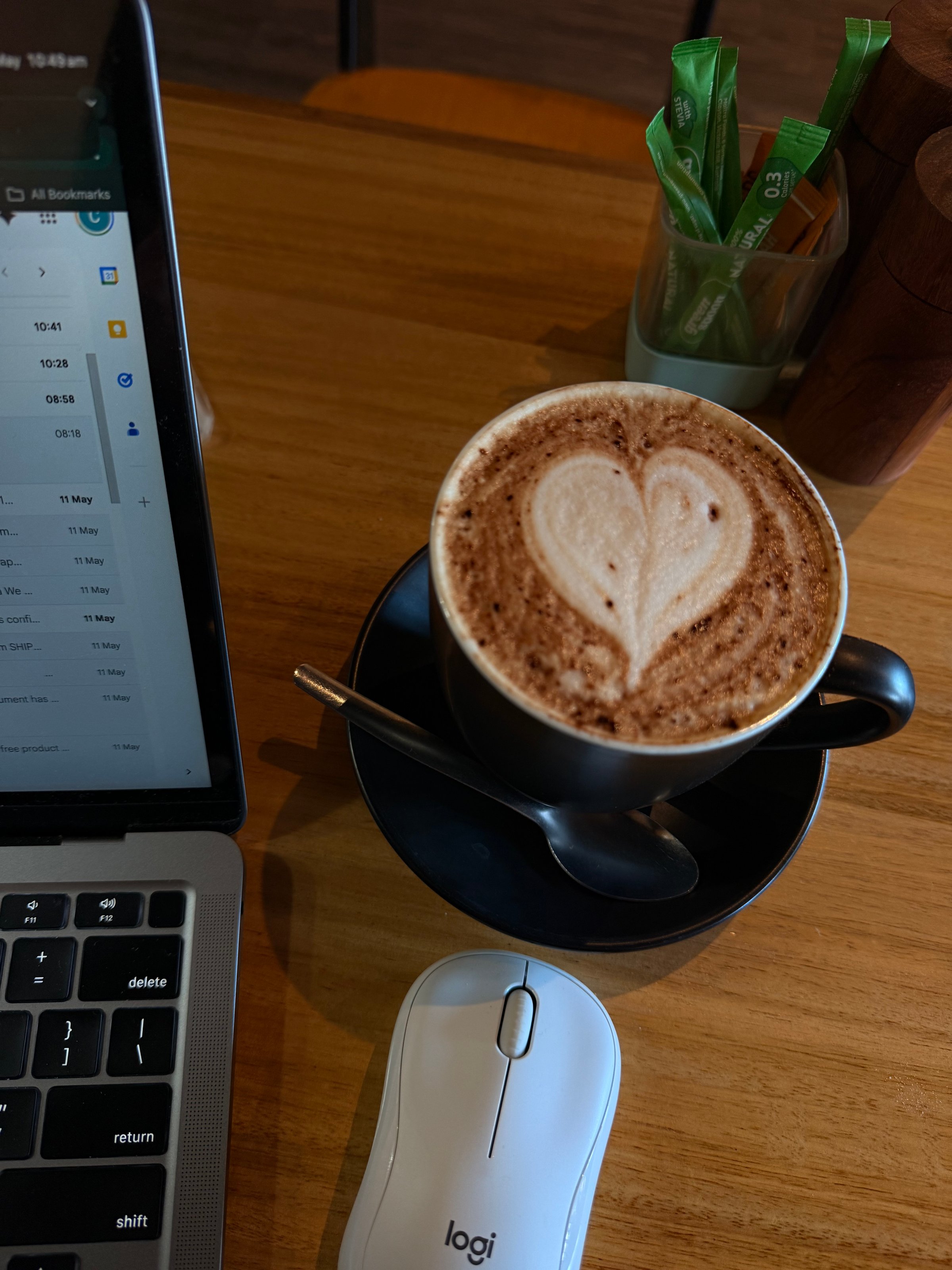 A cup of cappuccino with a heart-shaped foam design sits on a black saucer next to a laptop and a computer mouse on a wooden table.