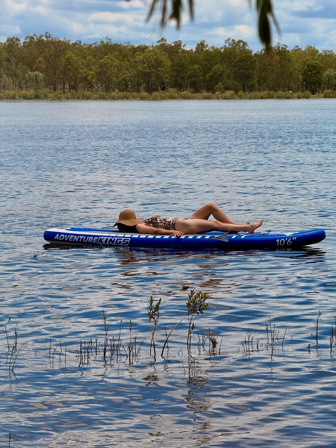 A person relaxing on a blue paddle board in a serene lake surrounded by greenery under a cloudy sky.