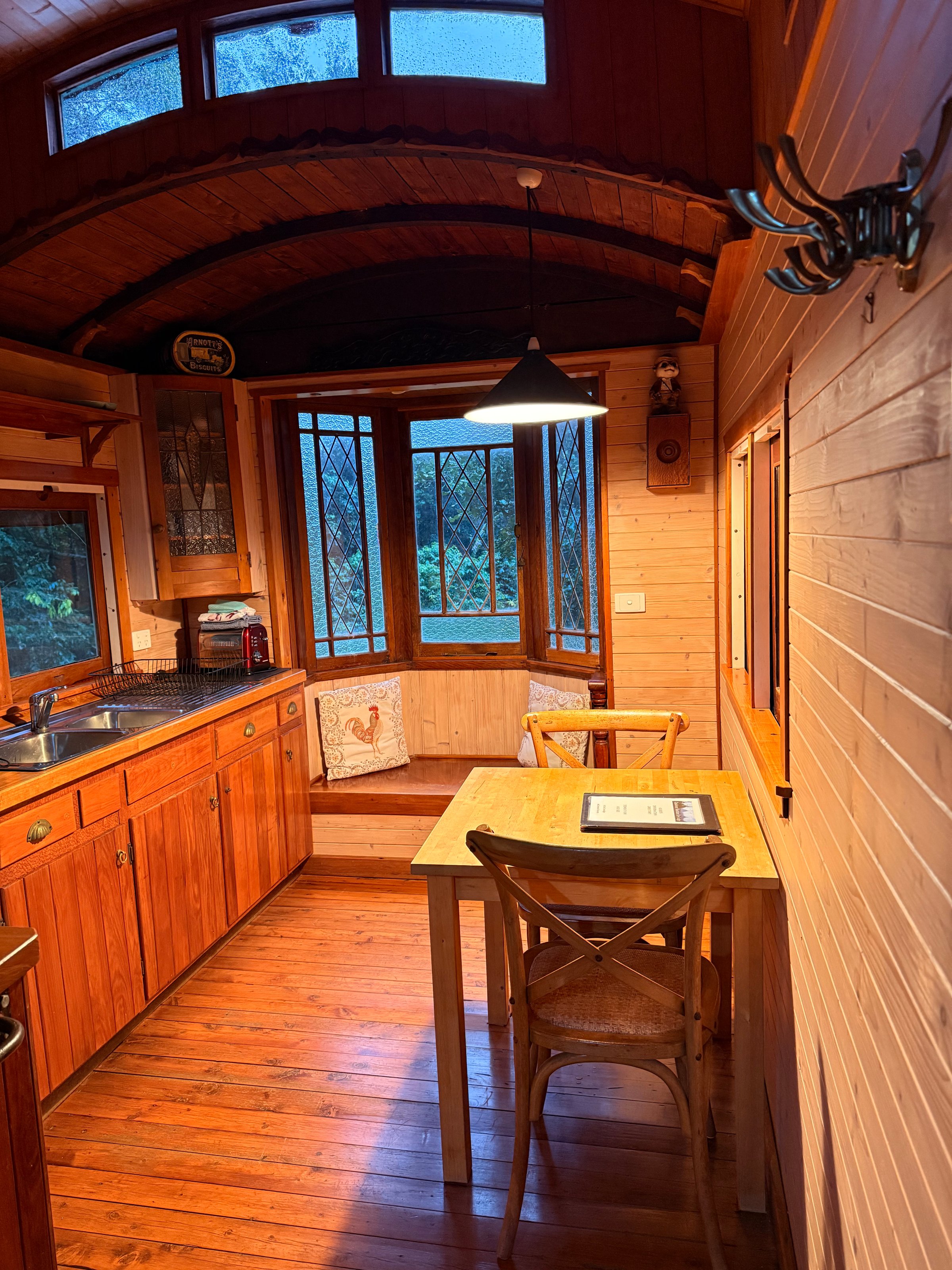 A cozy wooden kitchen with a table and chairs, featuring large windows that let in natural light and showcase a view of greenery outside.