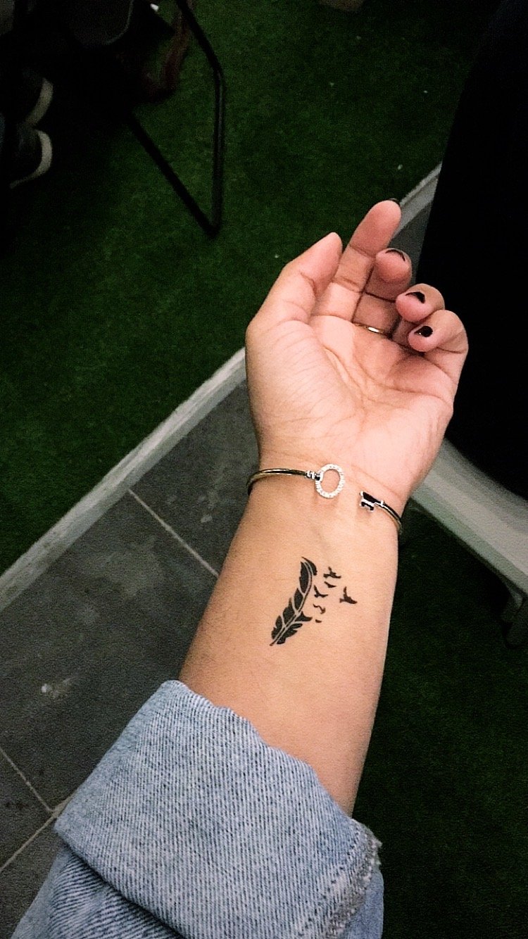 A close-up of a hand with a tattoo of a feather on the wrist, adorned with a silver bracelet.
