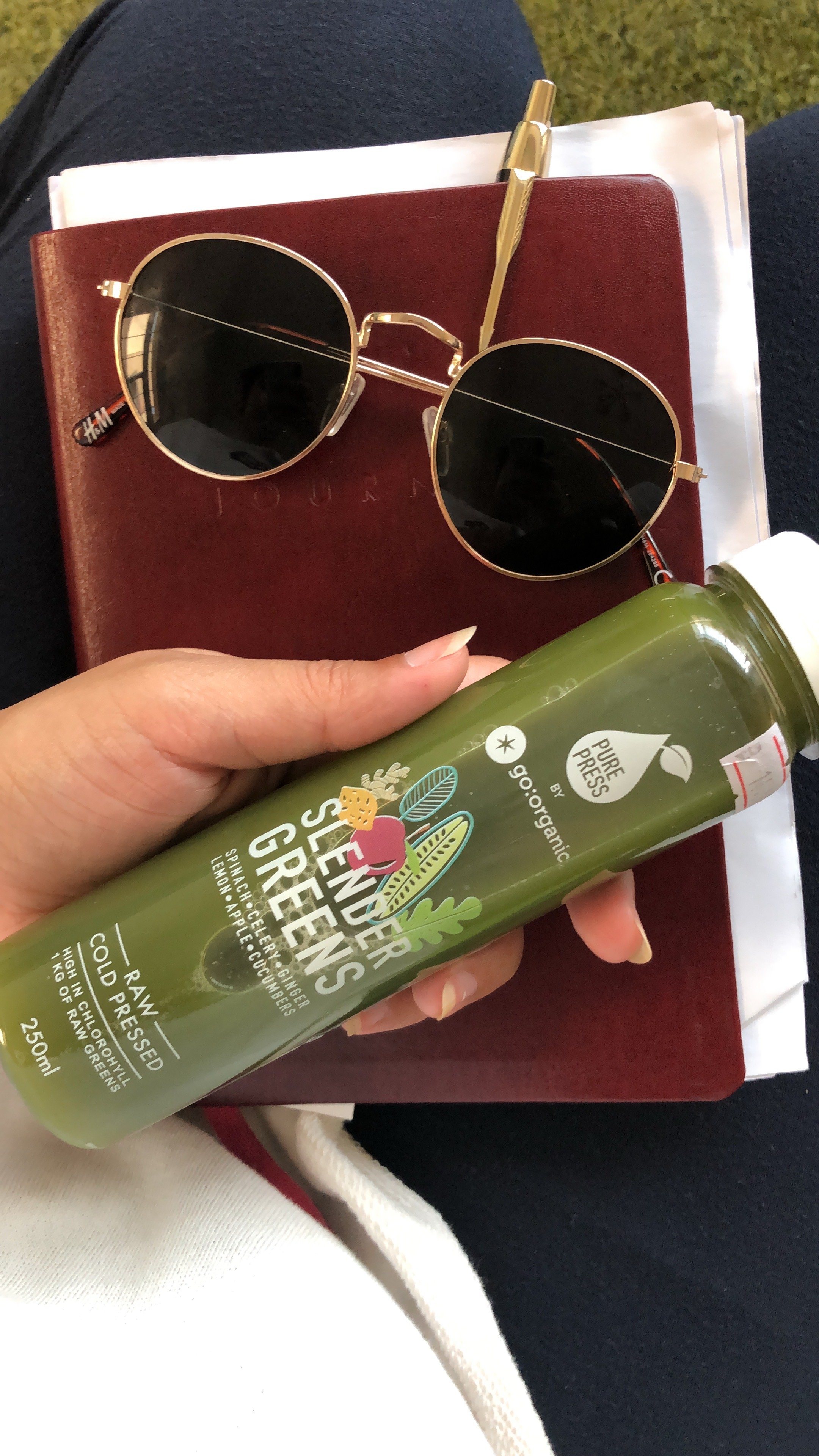 A hand holding a green juice bottle labeled 'Slender Greens' next to a pair of black sunglasses and a red notebook.