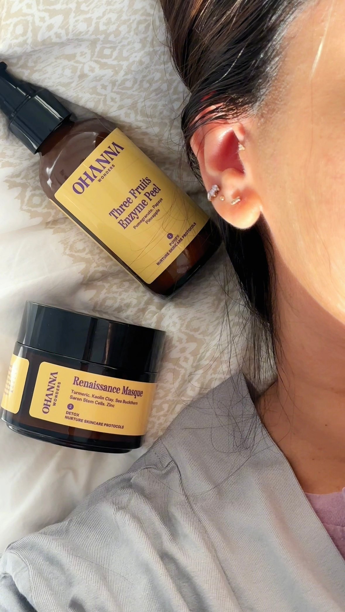 A close-up of skincare products placed near a person's ear, featuring a bottle of enzyme peel and a jar of mask.