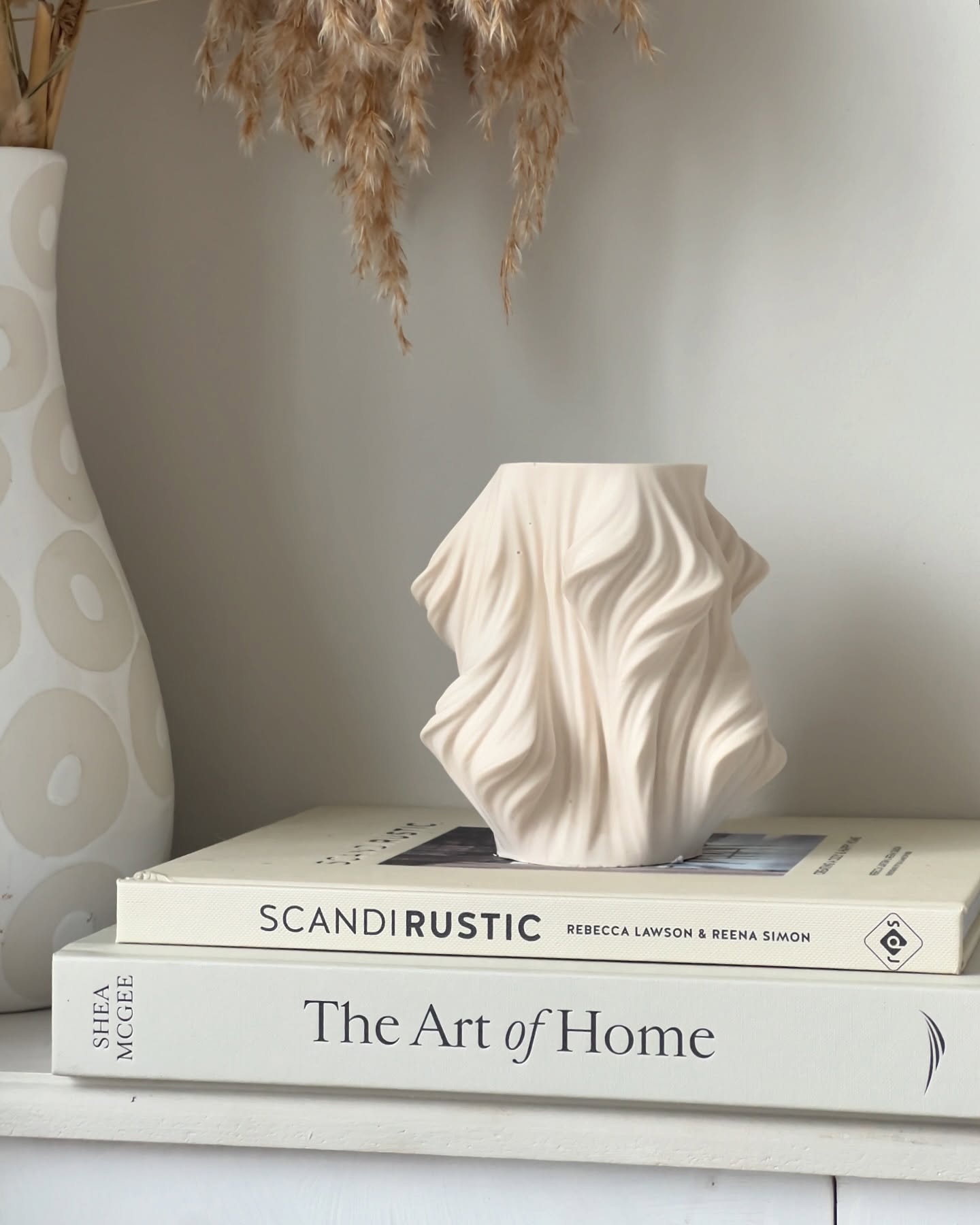 A textured beige vase with wavy patterns sits atop two stacked books, one titled 'SCANDIRUSTIC' and the other 'The Art of Home', with dried flowers in the background.