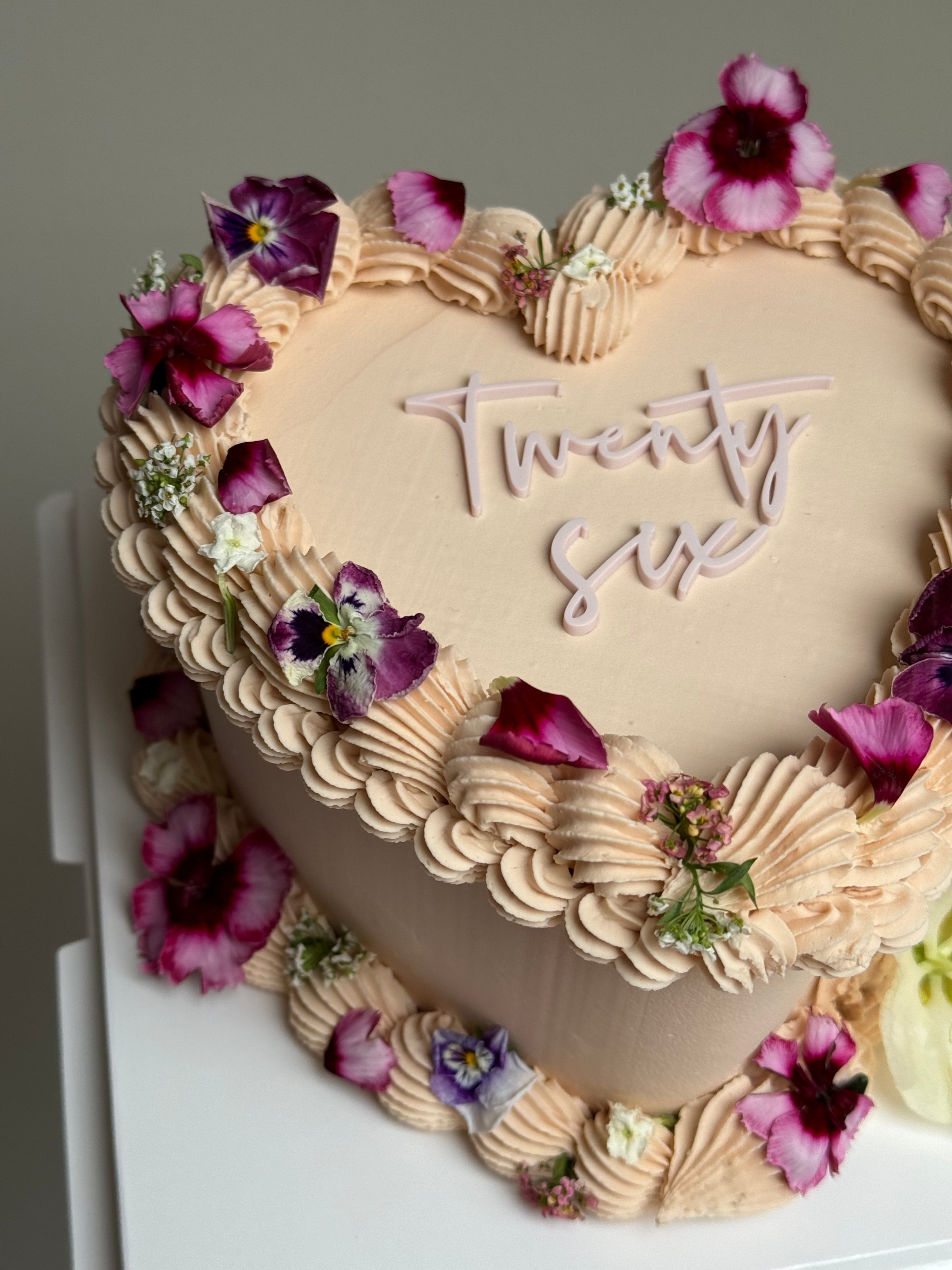 A heart-shaped cake decorated with pink frosting and edible flowers, featuring the inscription 'Twenty Six' in elegant script.