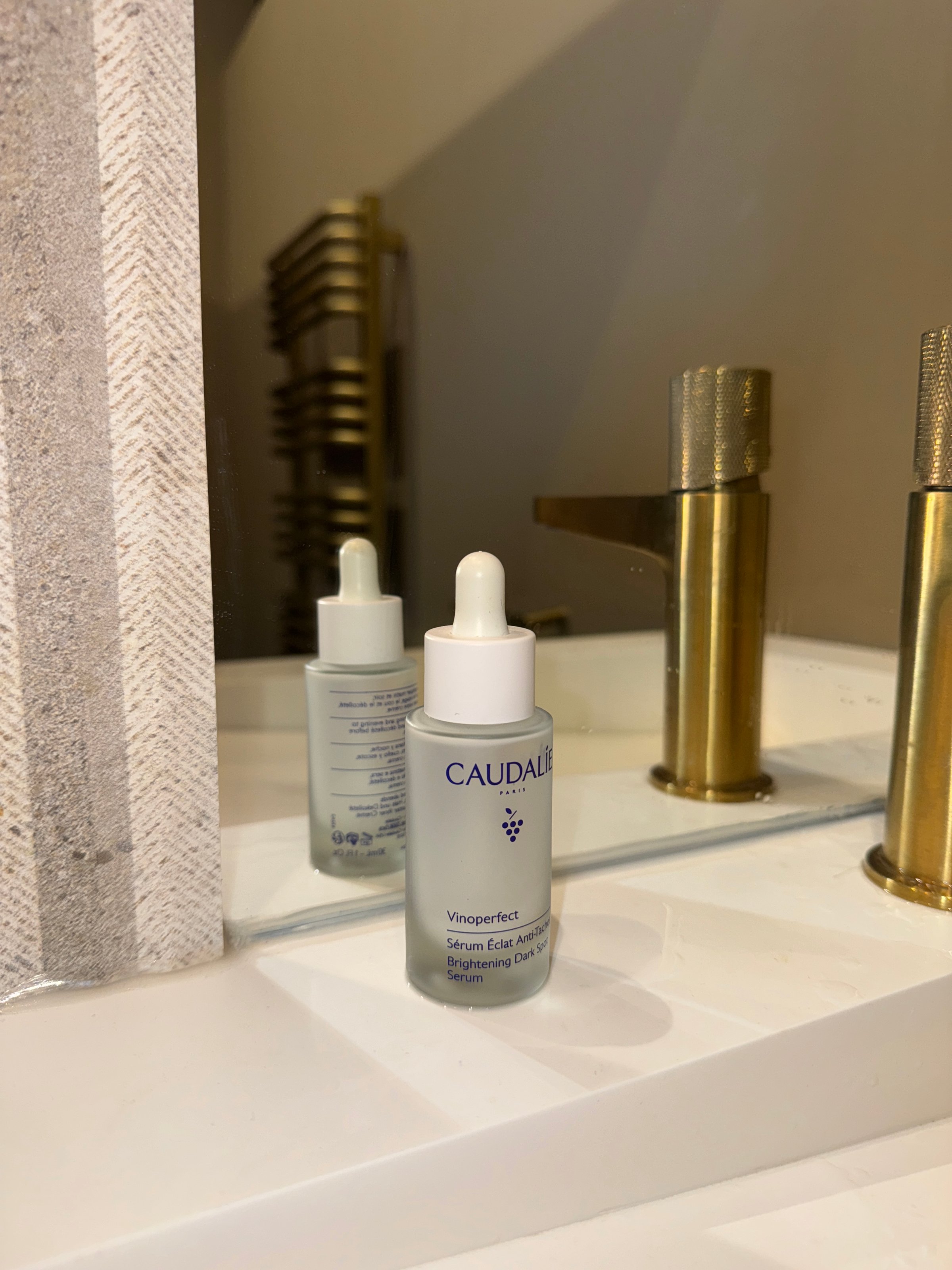 A bottle of Caudalie Vinoperfect serum is placed on a bathroom counter, reflecting in the mirror. The setting features a modern faucet and textured wall.