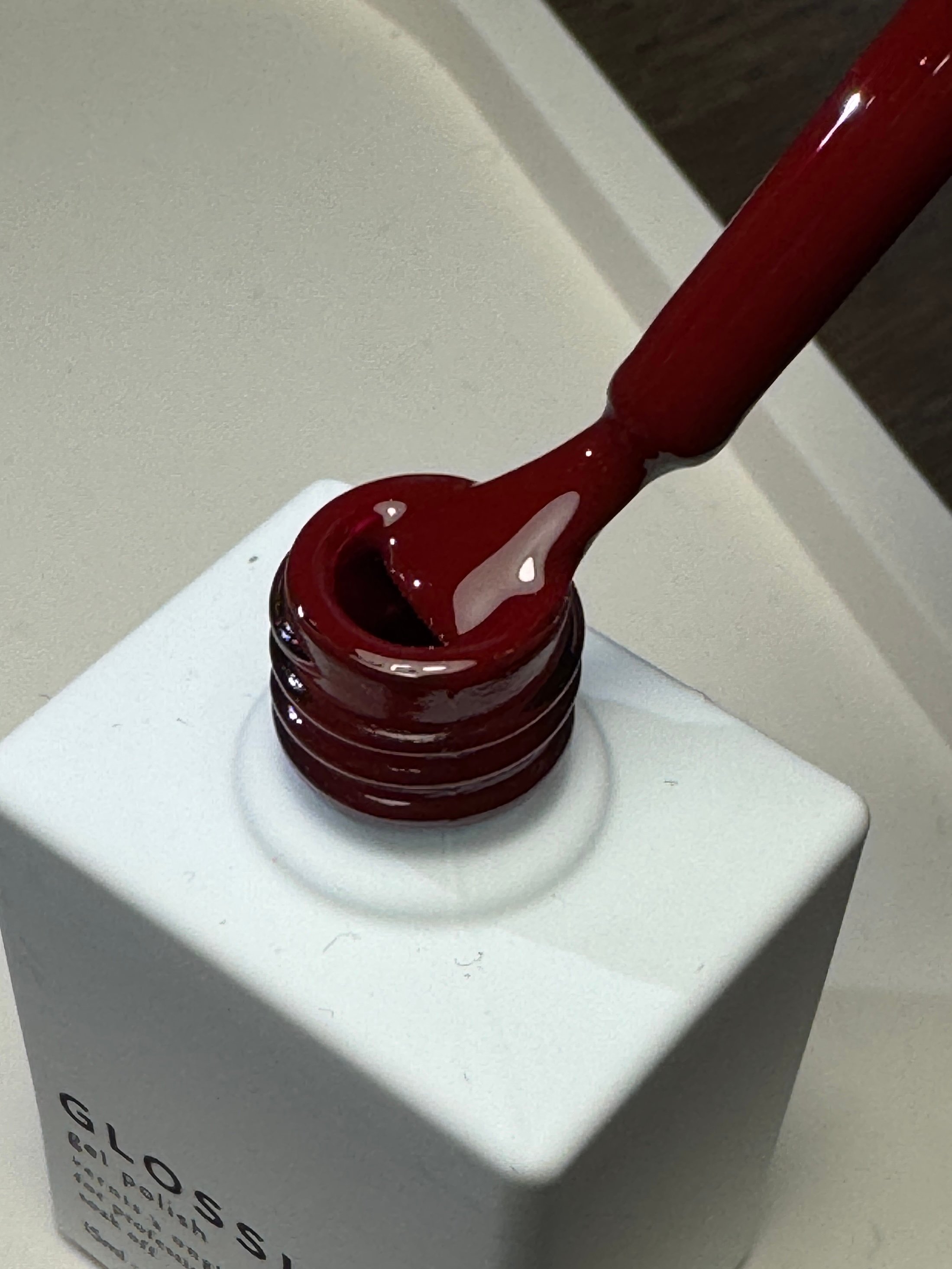 A close-up of a glossy red nail polish being pulled from a white bottle, showcasing the rich color and smooth texture of the polish.