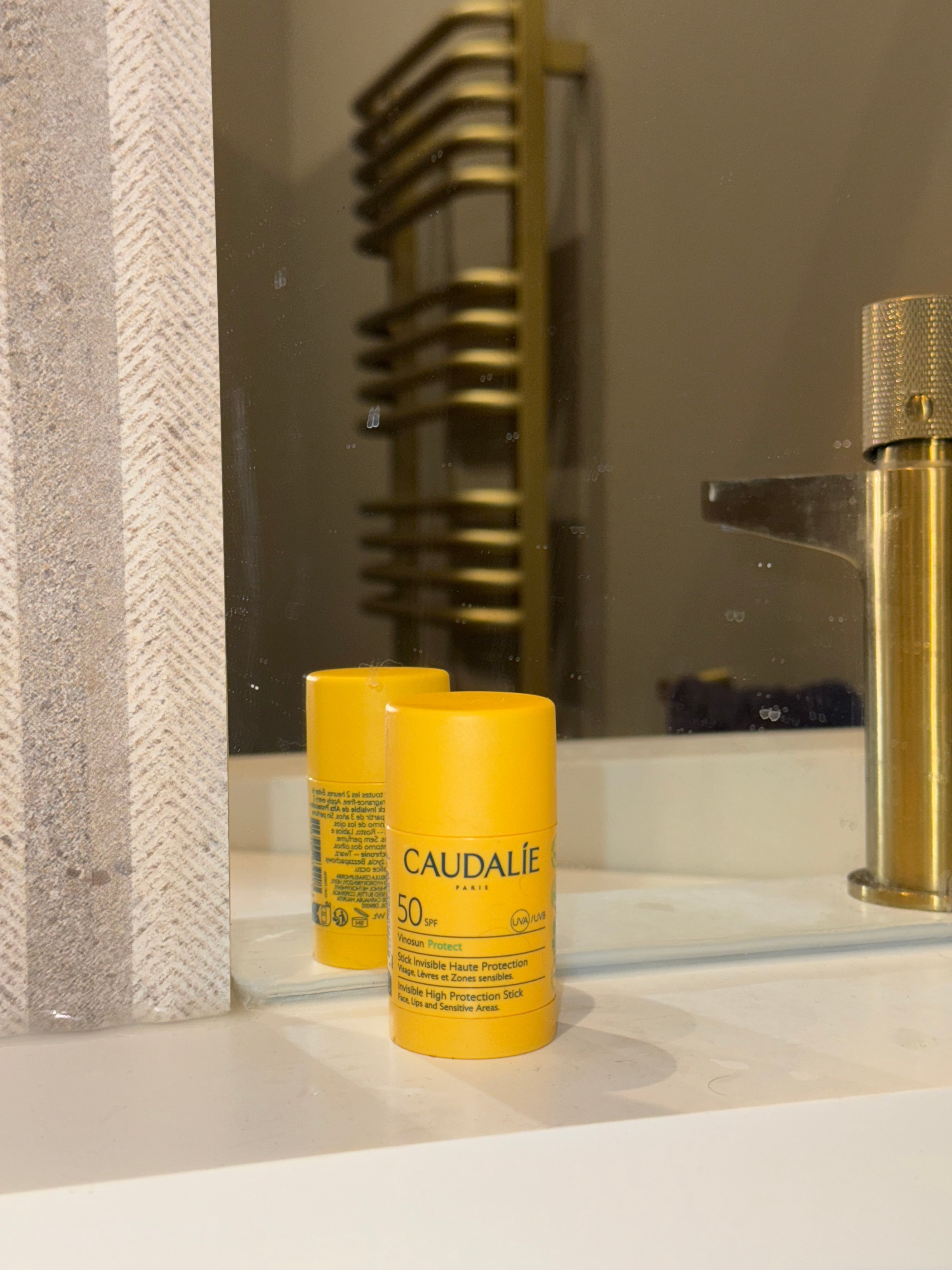 A yellow sunscreen stick from Caudalie is placed on a bathroom countertop, reflecting in the mirror behind it.