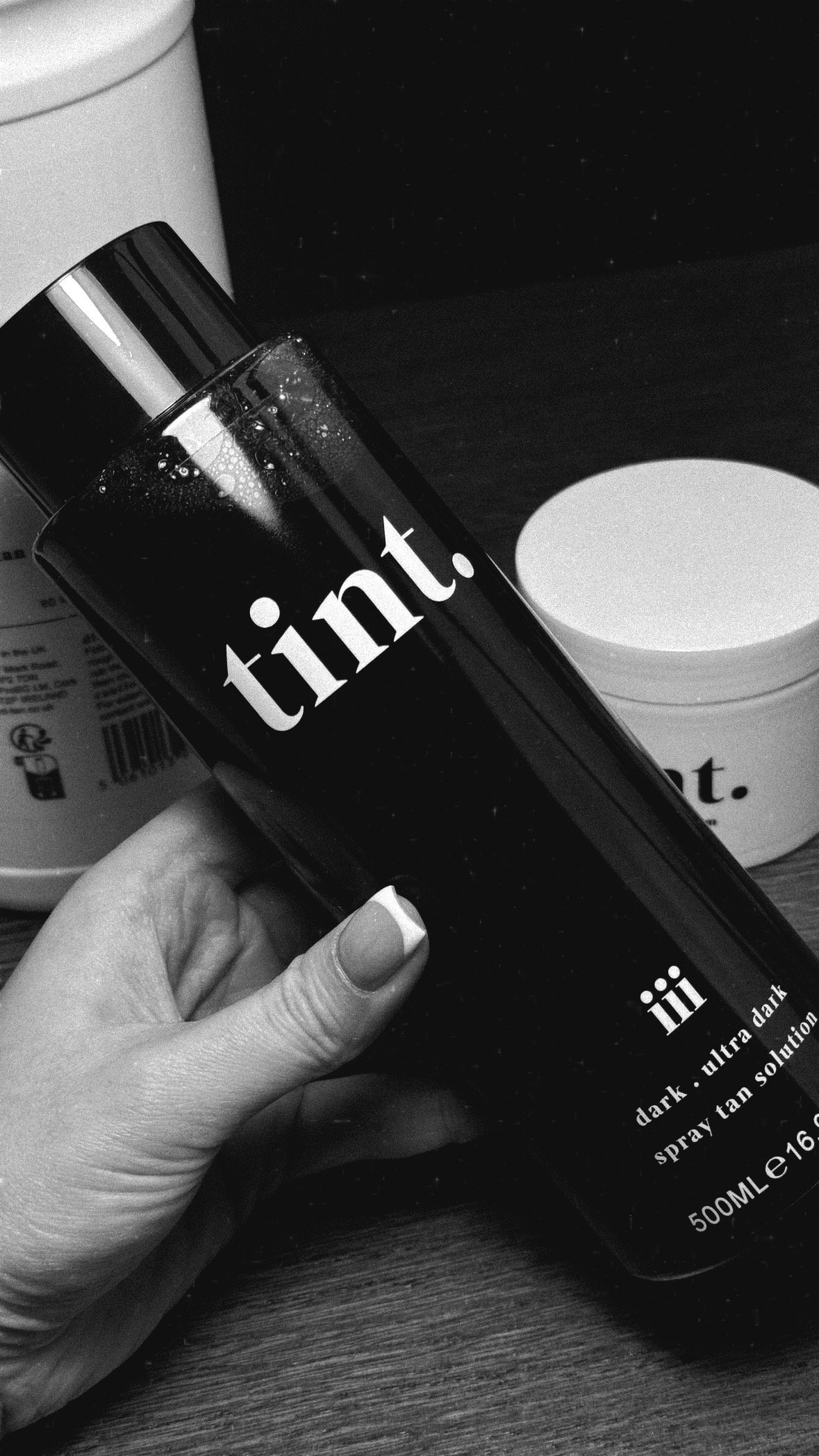 A hand holding a bottle of tint spray solution with a sleek black design and white text, placed on a wooden surface next to a white container.