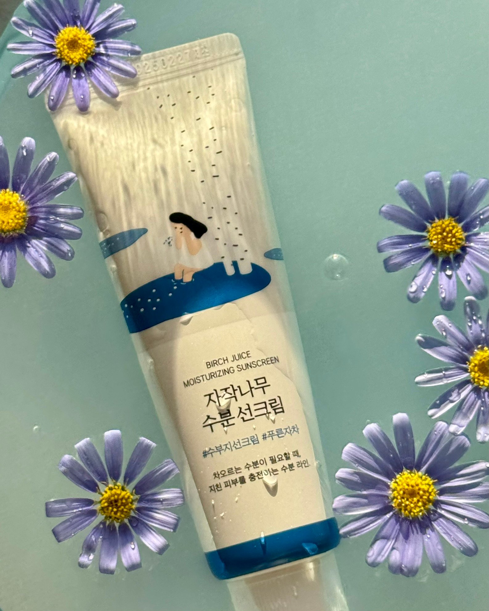 A tube of Birch Juice Moisturizing Sunscreen surrounded by purple flowers on a light blue background.
