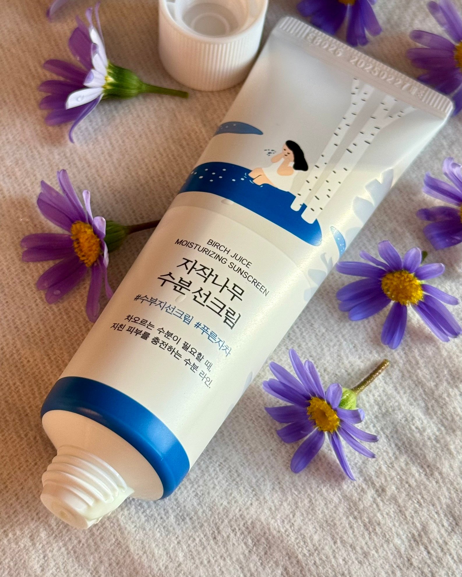 A tube of birch juice moisturizing sunscreen placed among purple flowers, showcasing its design and branding.