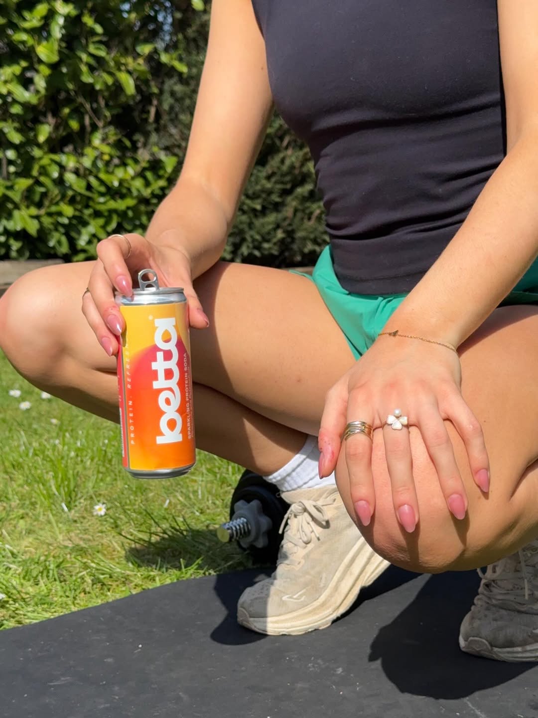A person squatting on a yoga mat in a garden, holding a can of Botta drink. They are wearing a black tank top and green shorts, with a focus on their hands adorned with rings and manicured nails.