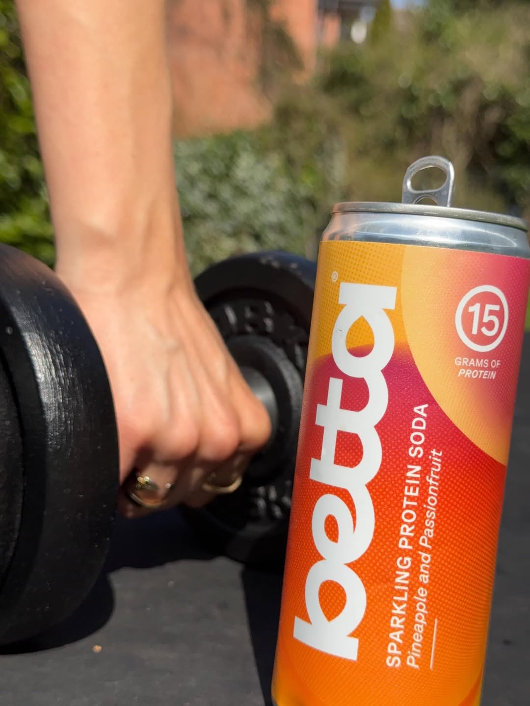 A hand holding a dumbbell next to a can of Betta sparkling protein soda in pineapple and passionfruit flavor.