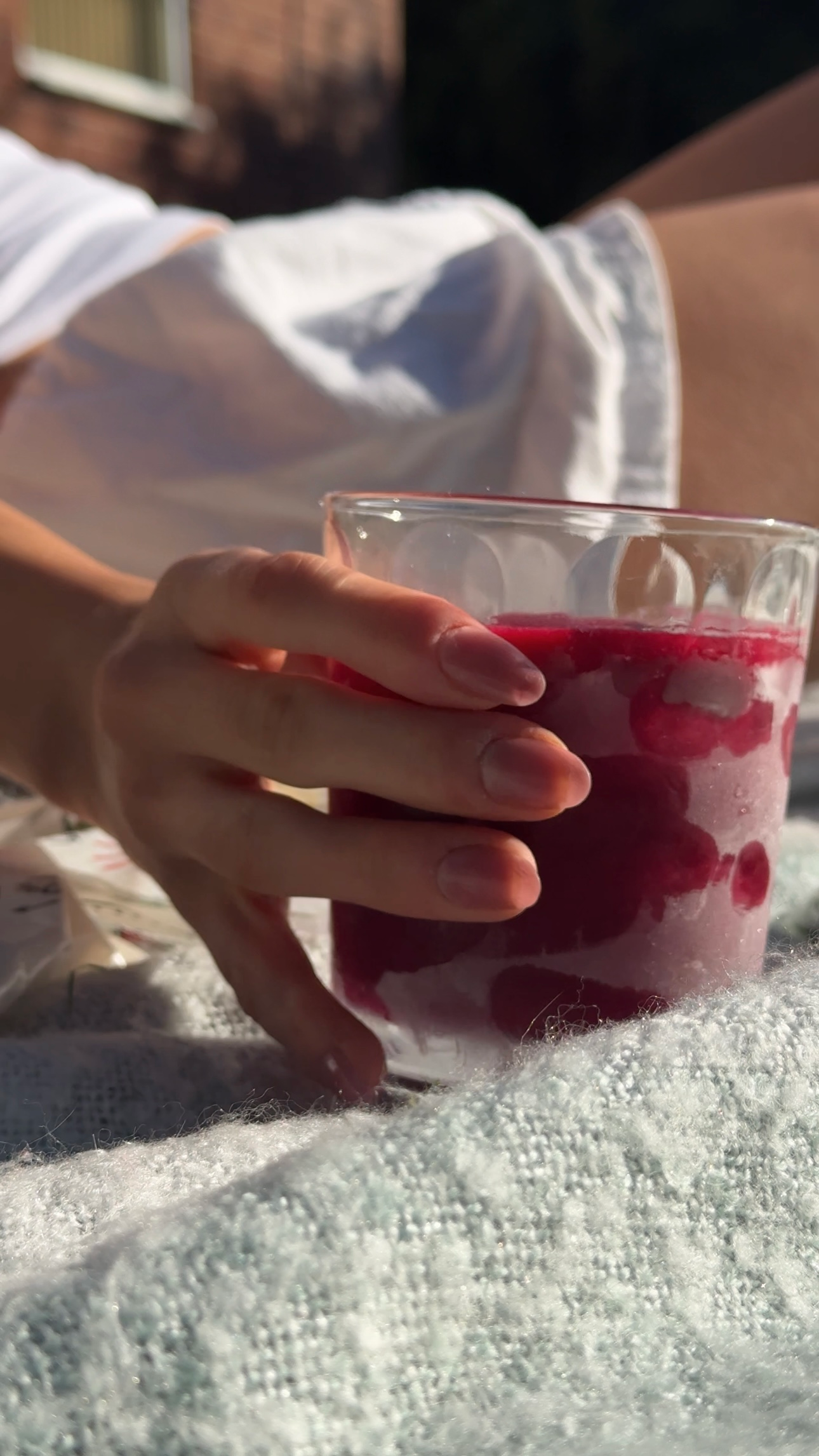 A close-up of a hand holding a glass filled with a red berry drink, resting on a soft, textured surface.