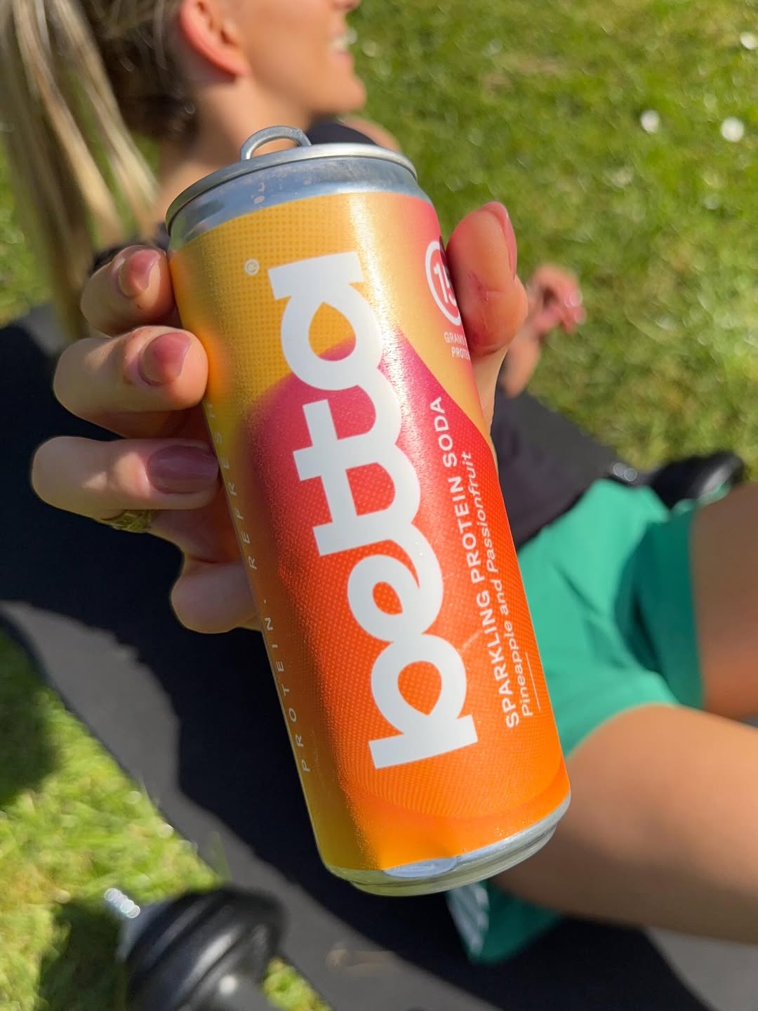 A person holding a can of sparkling protein soda in a vibrant orange and yellow design, with a blurred background of grass and a partially visible individual relaxing.
