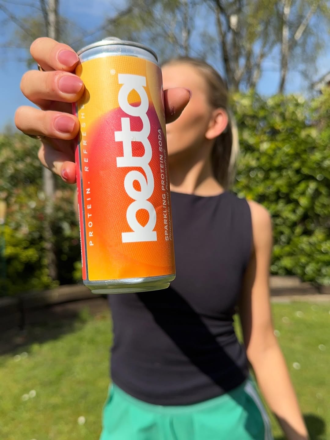 A person holding a can of Betta sparkling protein soda in a garden setting, with greenery in the background.
