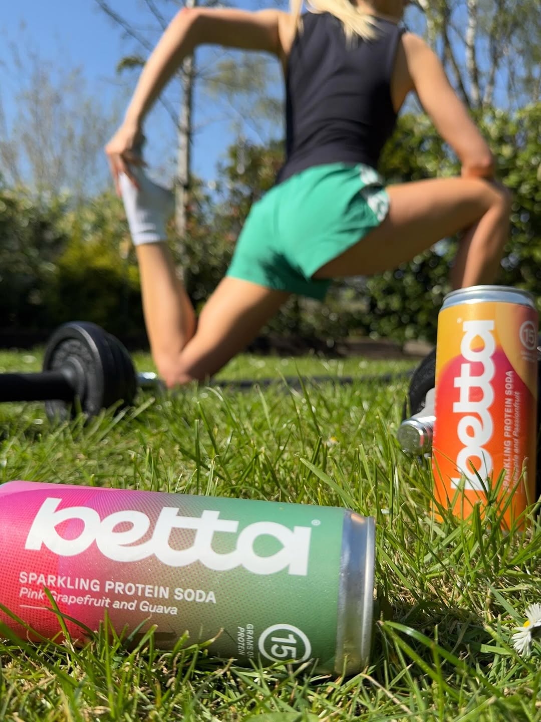 A person stretching in a grassy area with dumbbells in the foreground and cans of Betta sparkling protein soda nearby.
