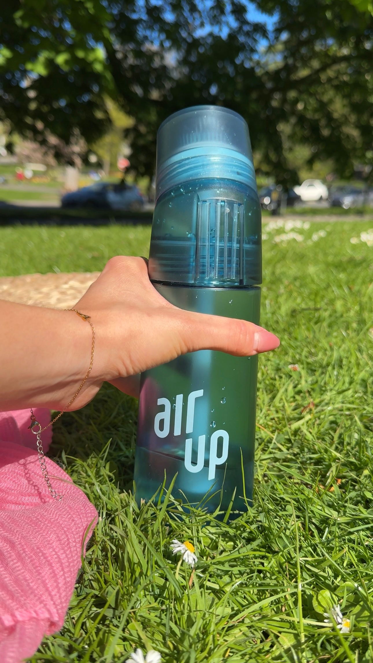 A hand holding a blue water bottle labeled 'air up' on a grassy field with a sunny background.