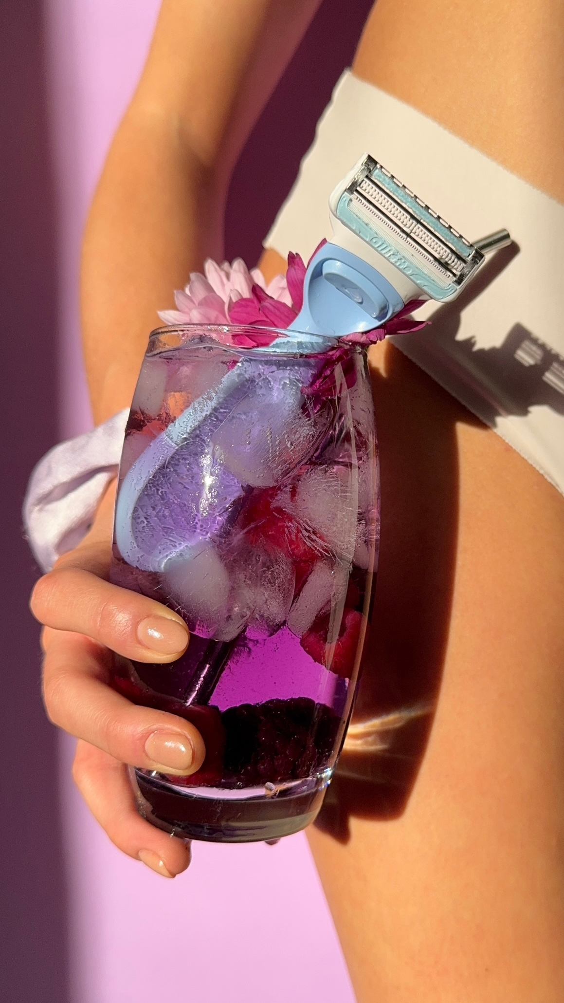 A hand holding a glass of colorful drink with ice and flowers, alongside a blue razor resting against the glass, set against a pastel pink background.