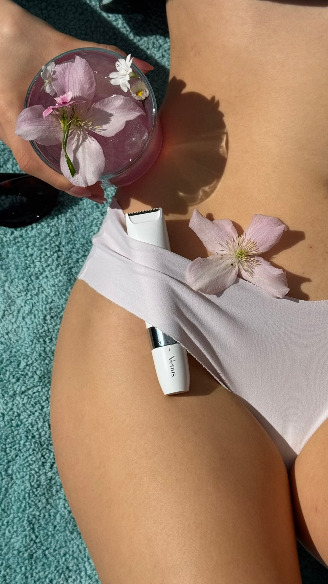 A close-up shot of a person's thigh adorned with pink flowers, alongside a glass filled with water and more flowers, set against a soft blue background.