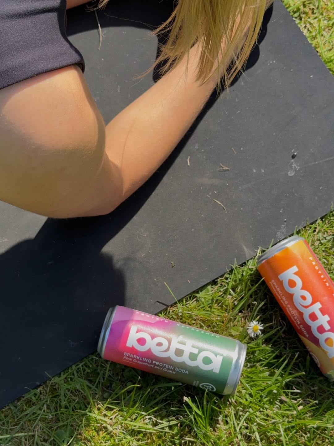 A close-up of a person's arm resting on a yoga mat, with two cans of sparkling protein soda placed on the grass beside them.