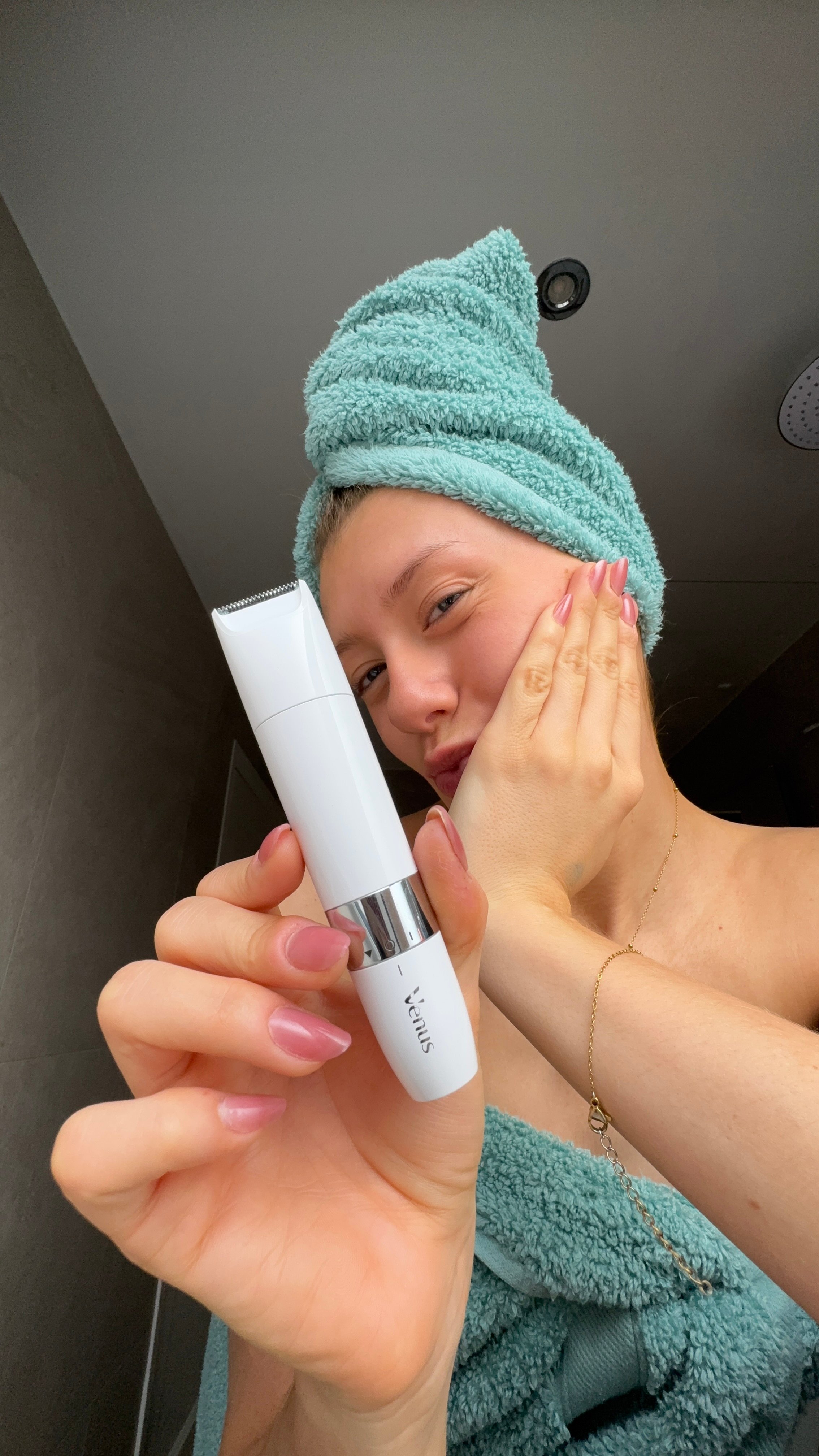 A person with a turquoise towel wrapped around their hair holds a skincare device while touching their face, showcasing a fresh and relaxed look.