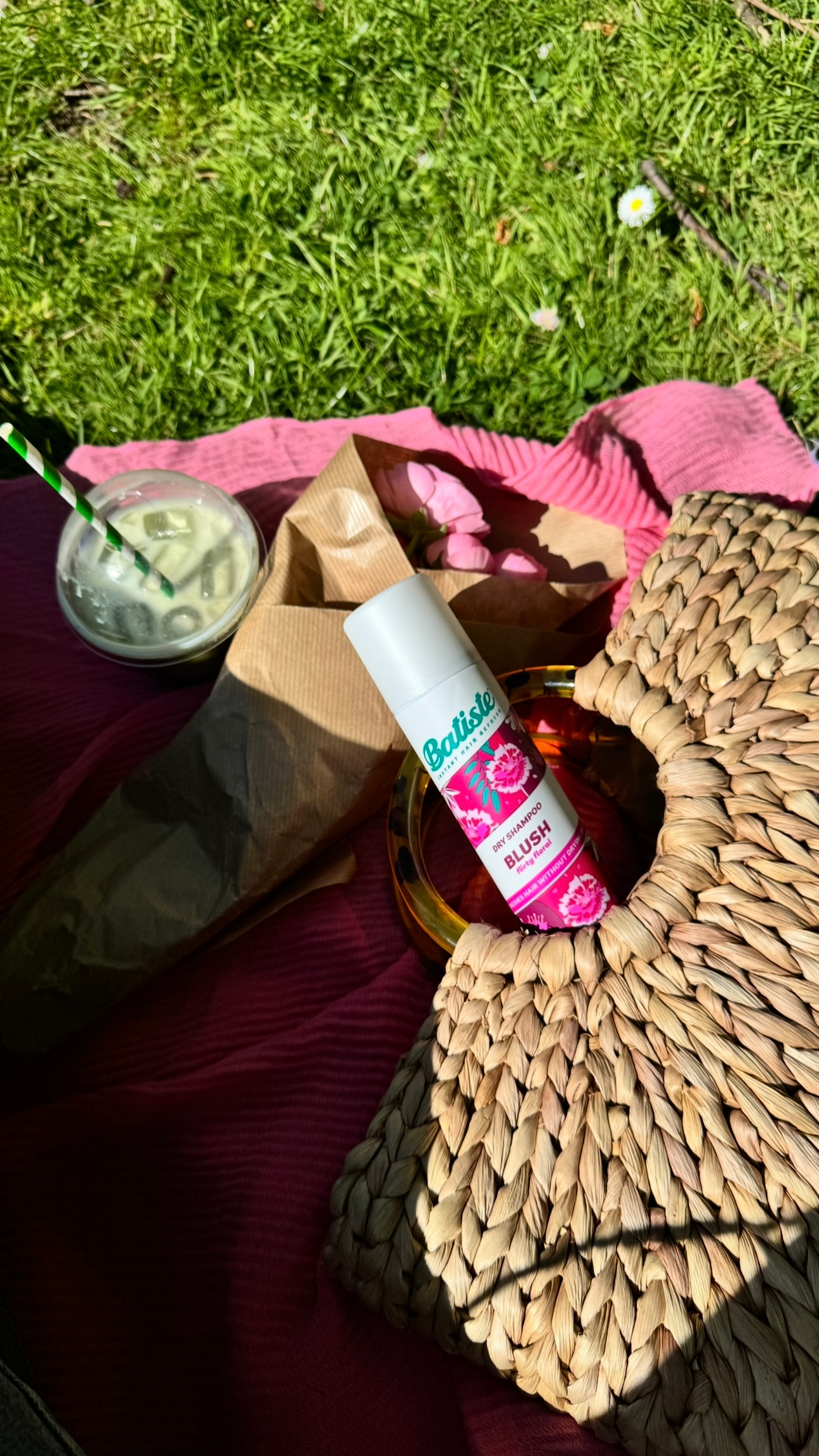A flat lay featuring a bottle of Batiste Blush dry shampoo, a woven bag, a wrapped snack, and a drink on a pink blanket in a grassy setting.