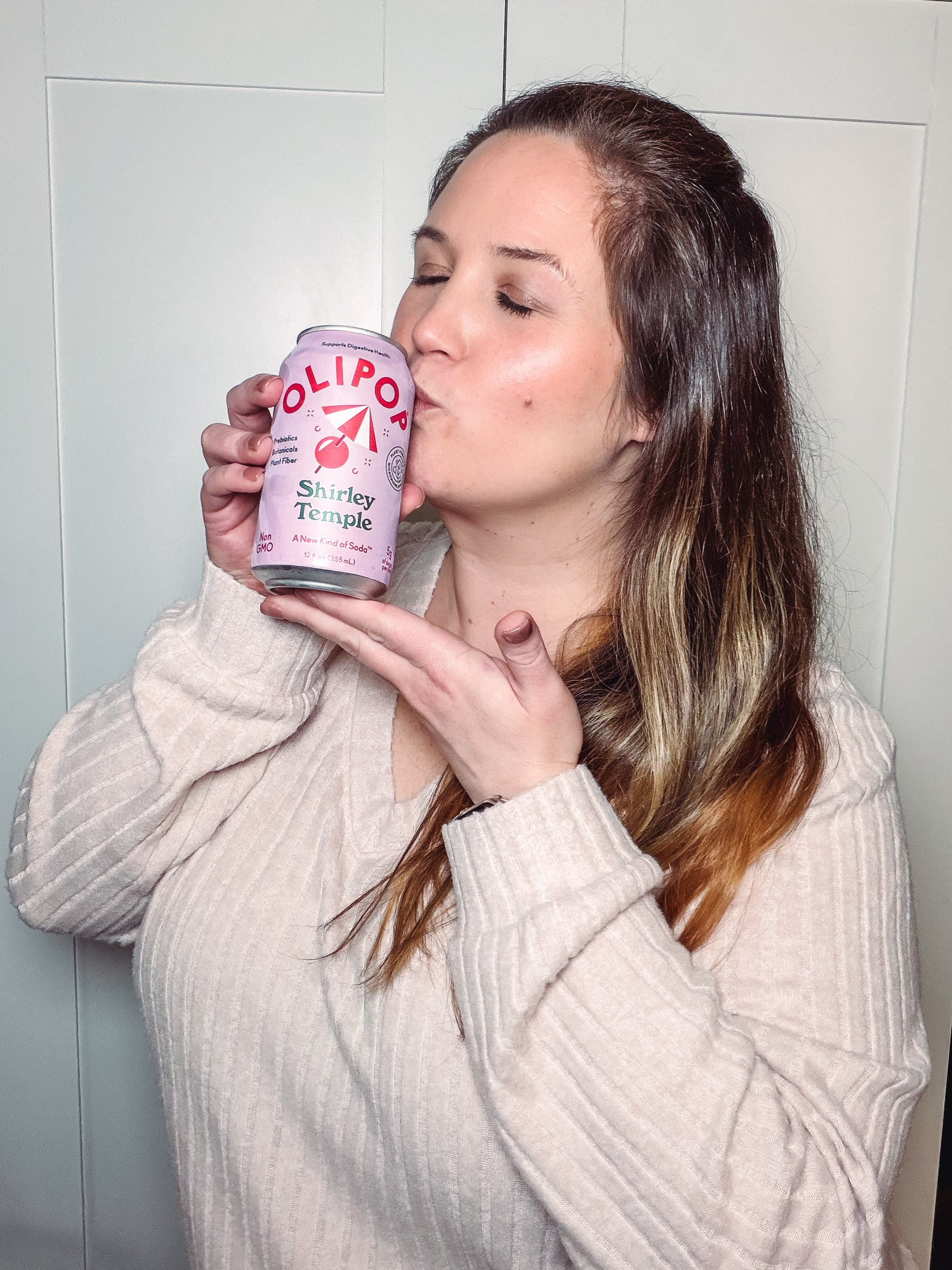 A woman in a cozy sweater kisses a can of Shirley Temple soda, showcasing a playful and joyful moment.