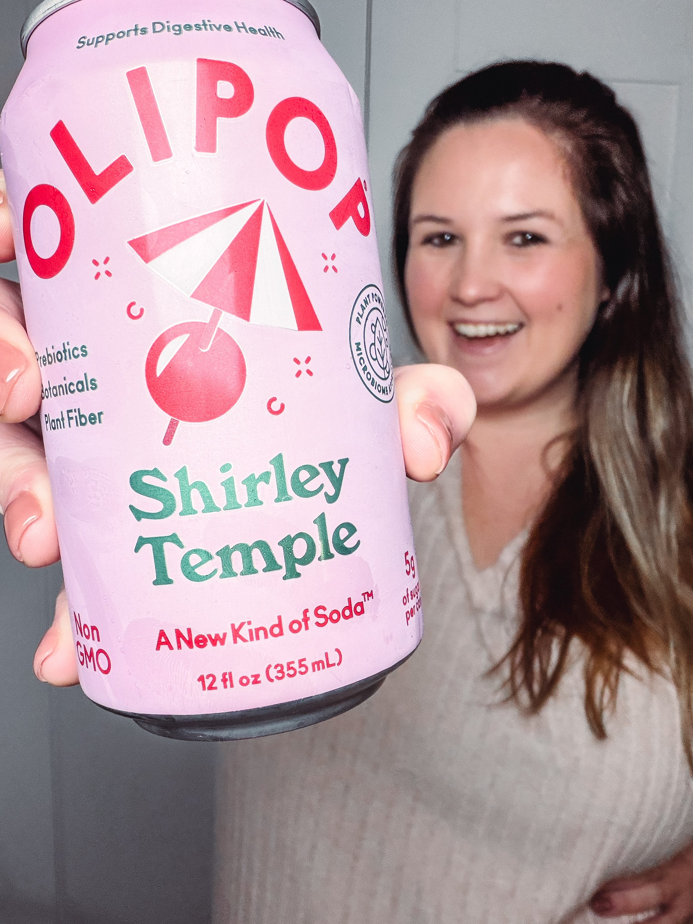 A person holding a can of OLIPOP Shirley Temple soda, smiling in the background. The can features a pink design with a cherry and an umbrella.
